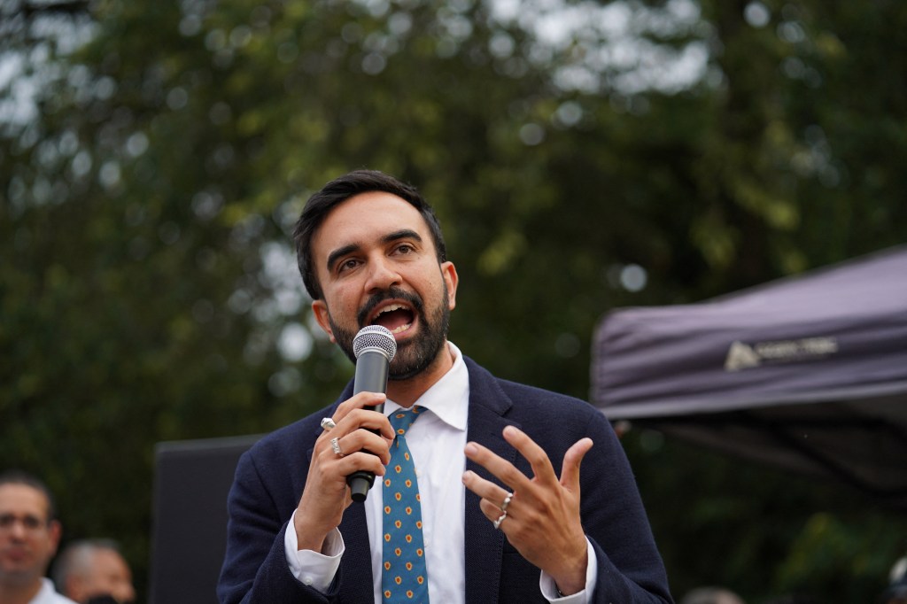 Zohran Mamdani speaking into a microphone at an outdoor event.