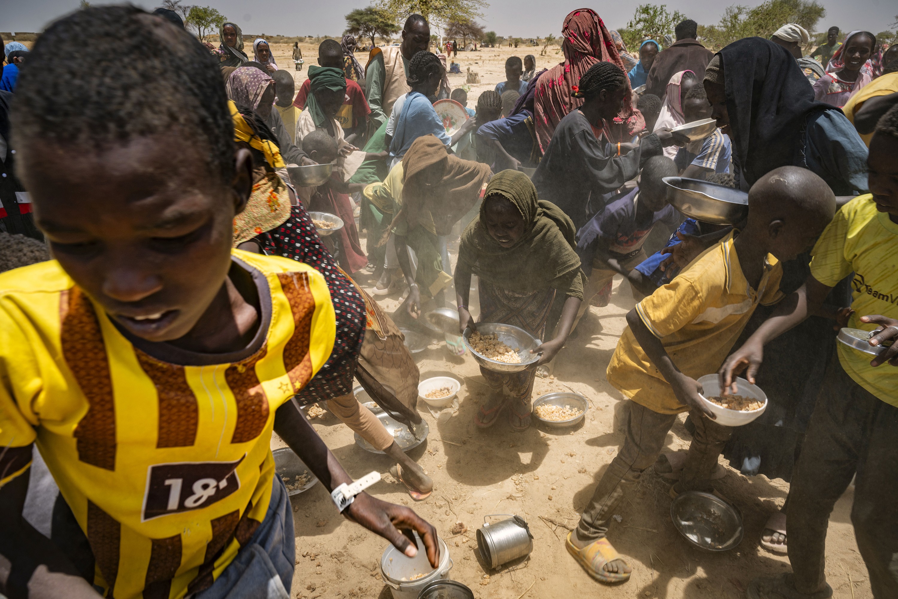 photo of food distribution with child wearing yellow soccer jersey in foreground and chaotic scene of women and children filling bowls with arid landscape in background