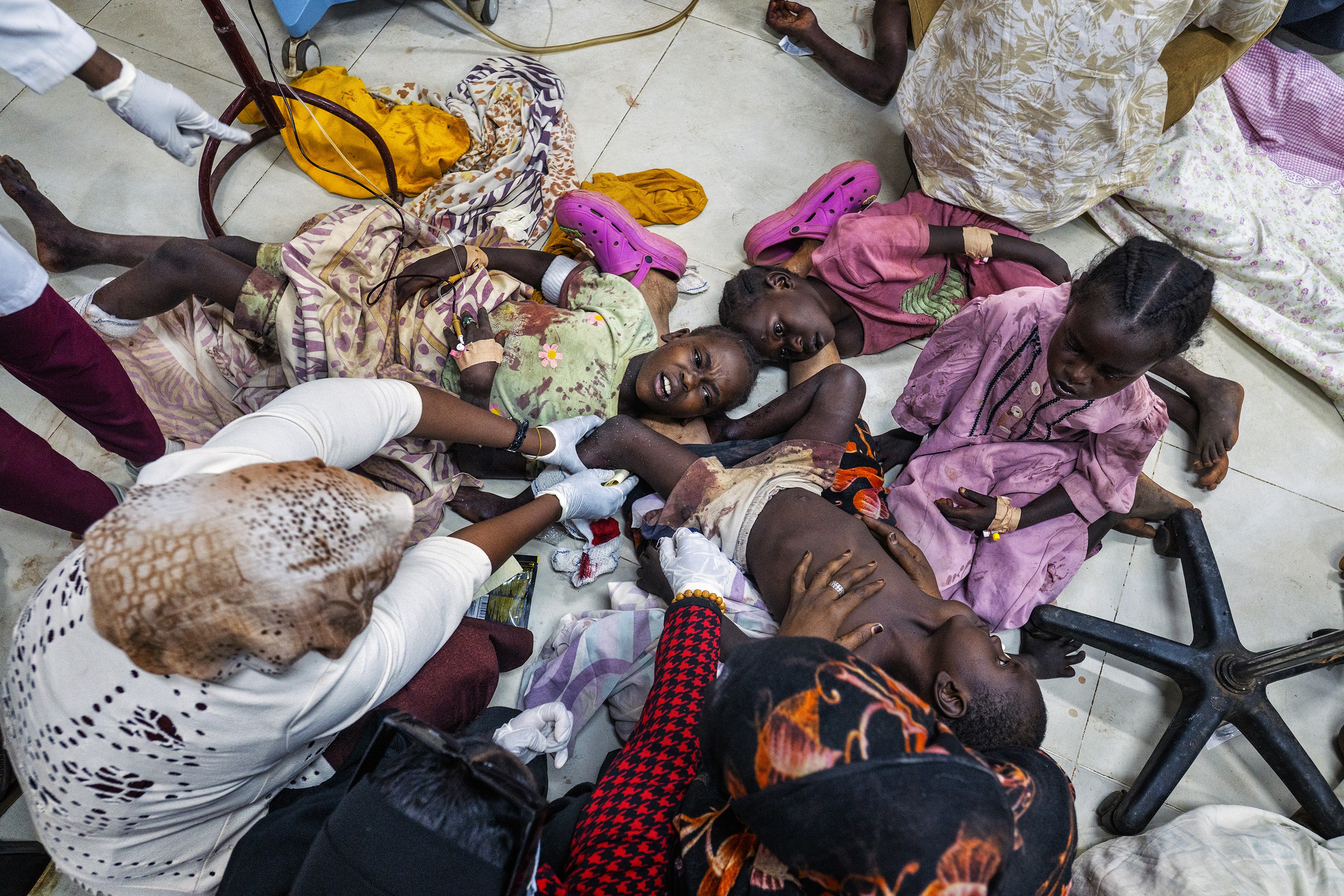 photo from overhead of nurses treating four small children lying in different positions on floor with limbs intertwined, two with their heads resting on a woman's outstretched legs wearing pink crocs and one held in her lap