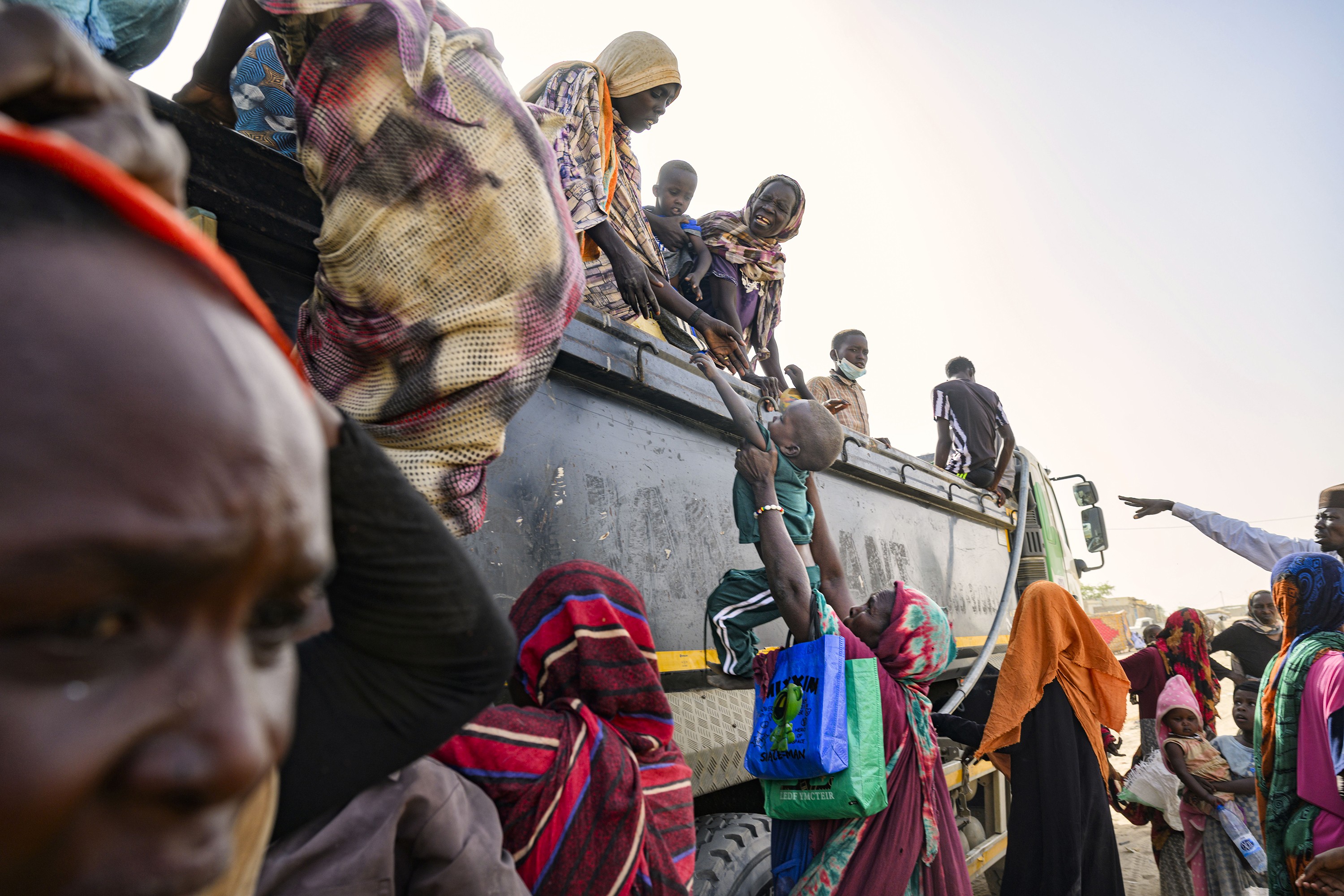 photo of people climbing into very tall-sided truck, with one woman on top holding a baby next to another woman reaching for a small child with arms outstretched that is being handed up to her from a woman on the ground, who is surrounded by other woman and children