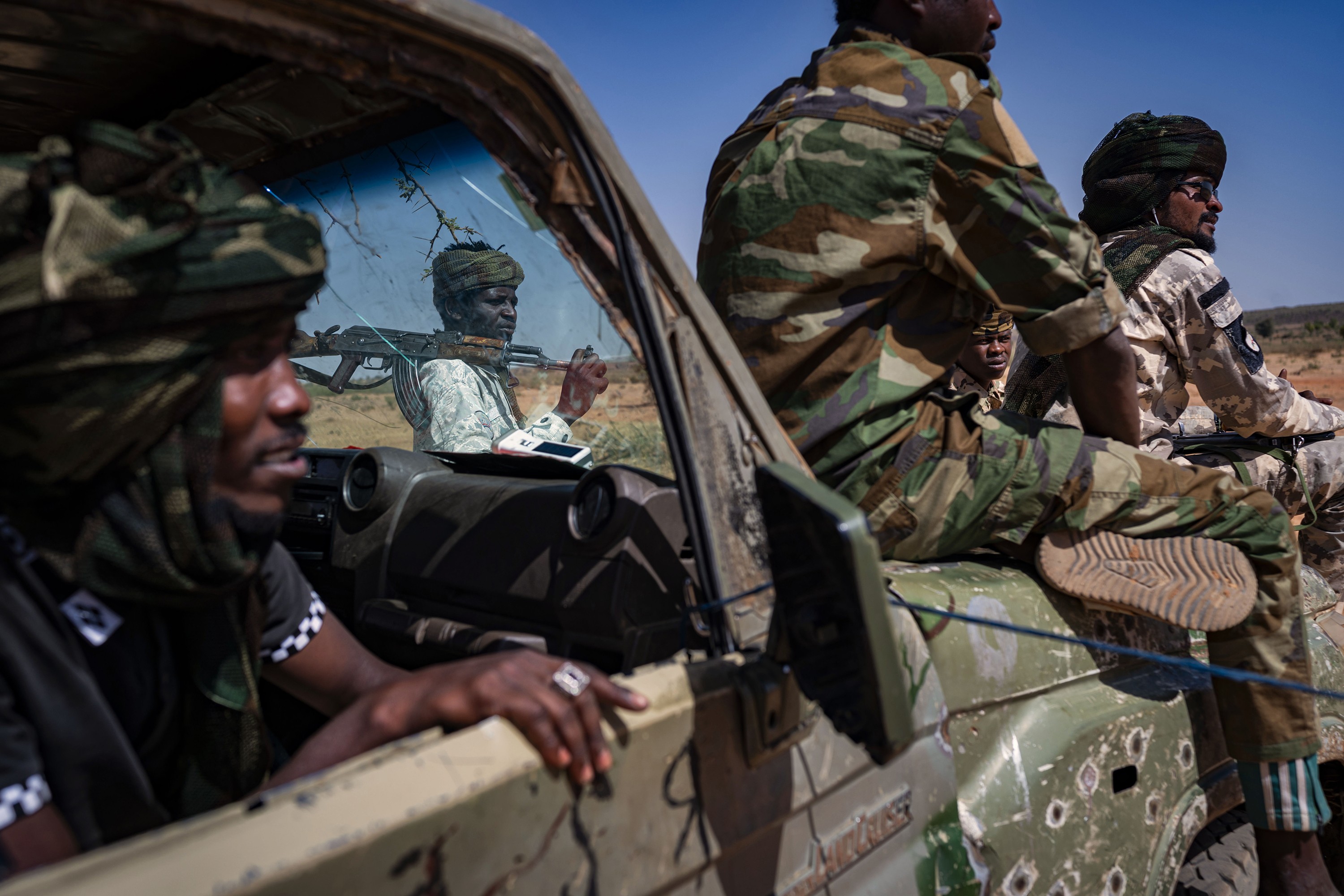 photo of right side of military truck pockmarked with bulletholes, with man inside leaning out open window and two more soldiers sitting on the hood, and through cracked windshield a third man visible standing with rifle over shoulder.