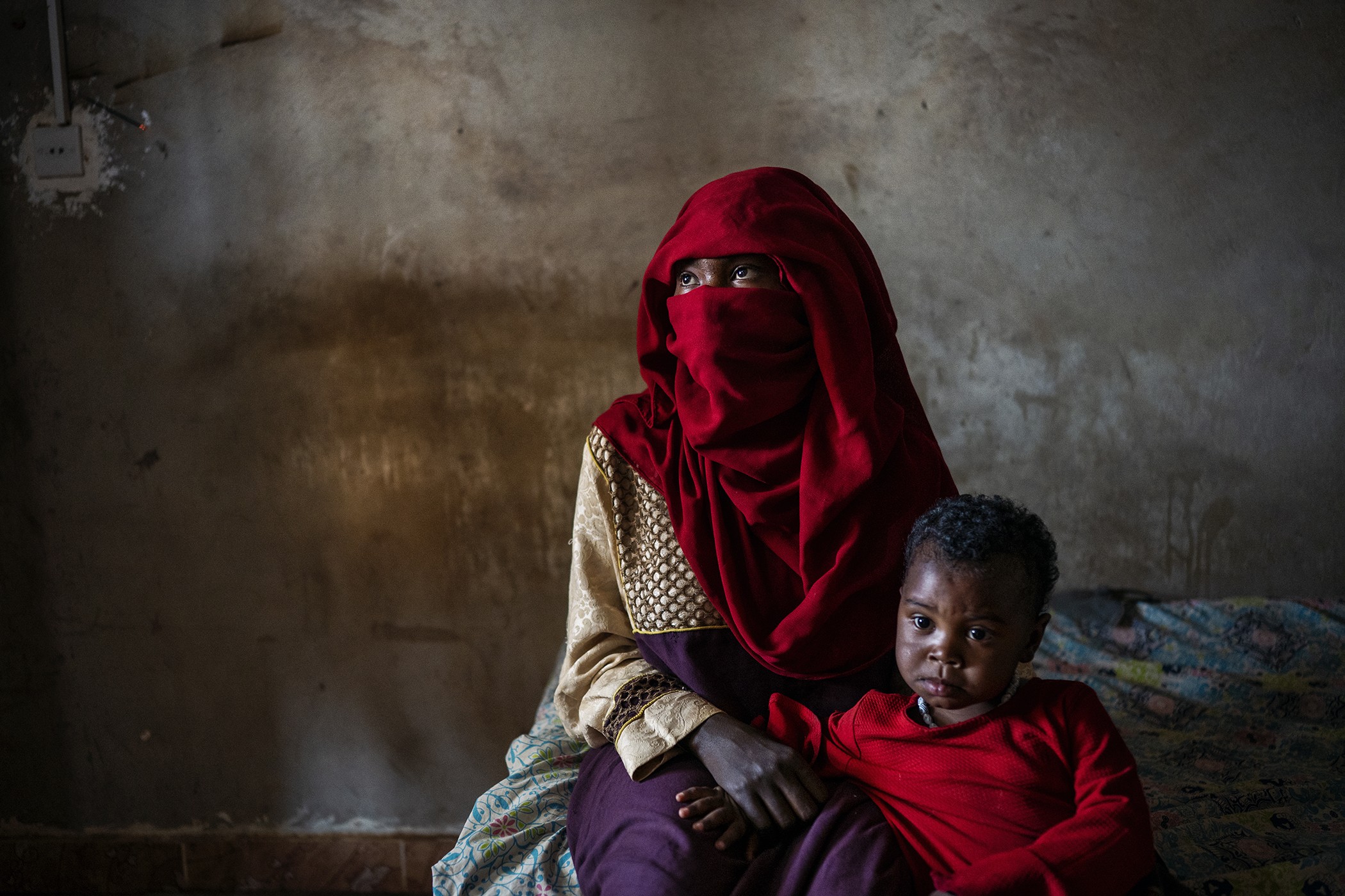 a woman in a brilliant-red head scarf and face covering sitting in room, looking up and to left, with young child sitting in her lap looking at camera