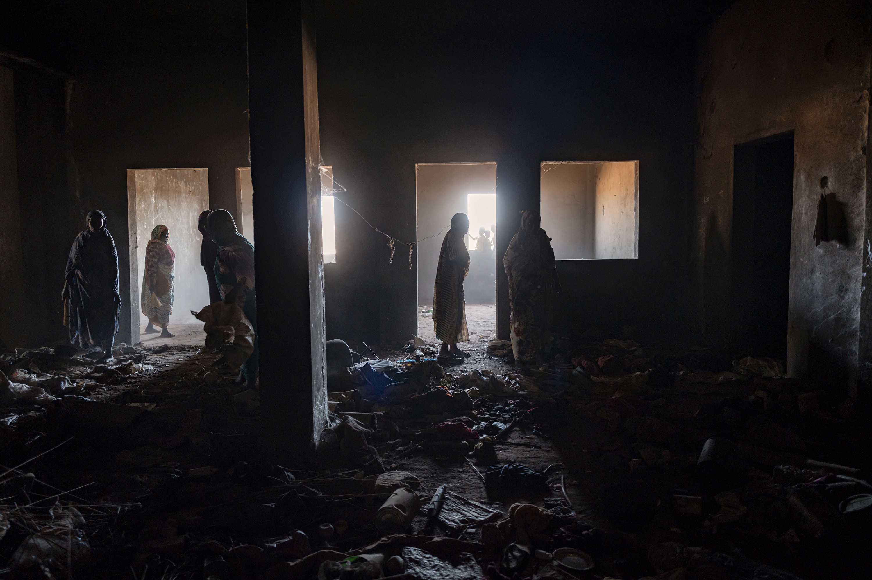 photo of the dark interior of a debris-strewn room, with numerous people silhouetted in light coming through open doorways in the background