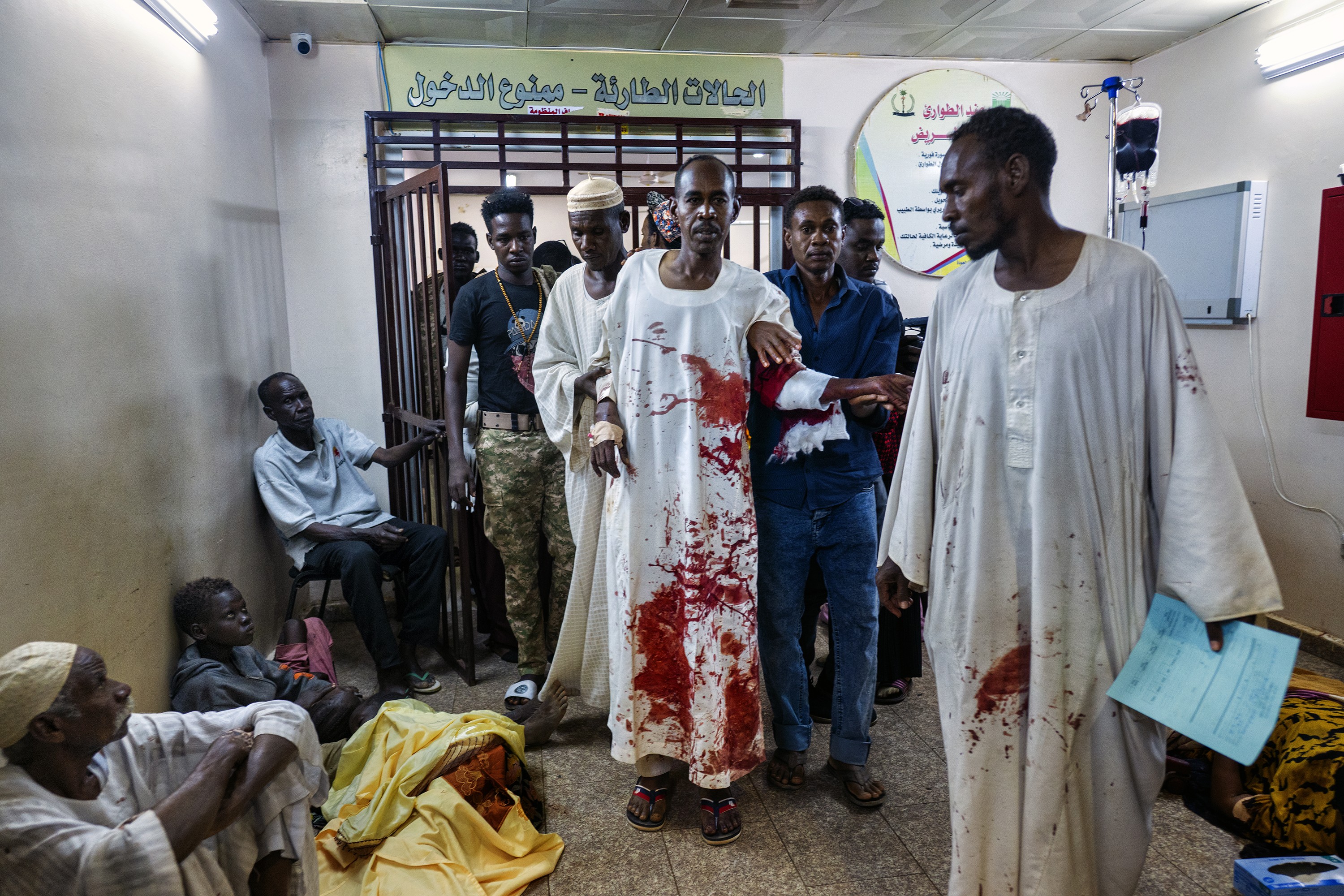 photo of group of men entering iron-gated doorway with door open, two of them helping a third walking in long white robe covered in bright crimson blood, with more patients sitting along the wall