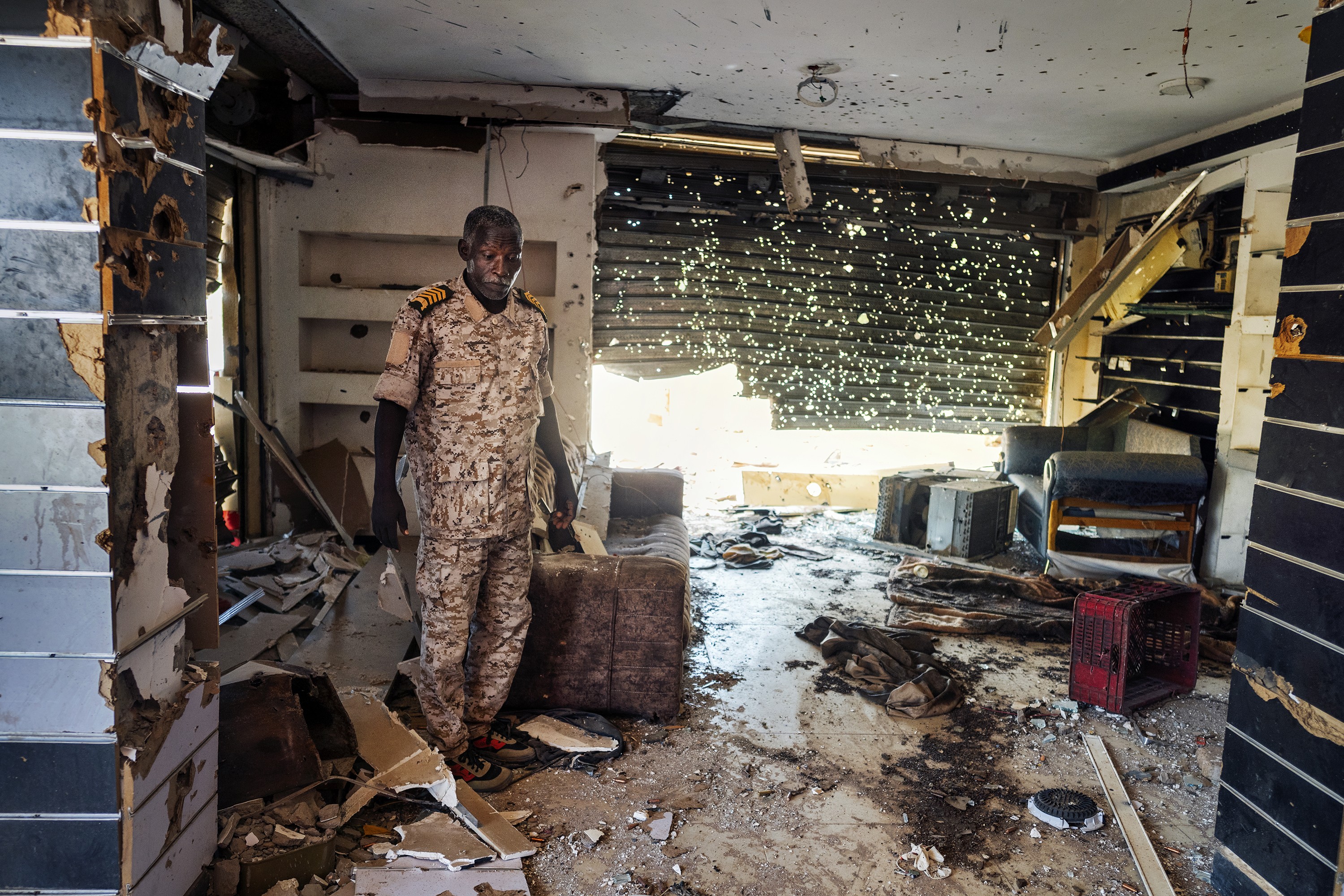 photo of man in military uniform in damaged room filled with debris, with roll-down door half-destroyed and riddled with bullet holes 