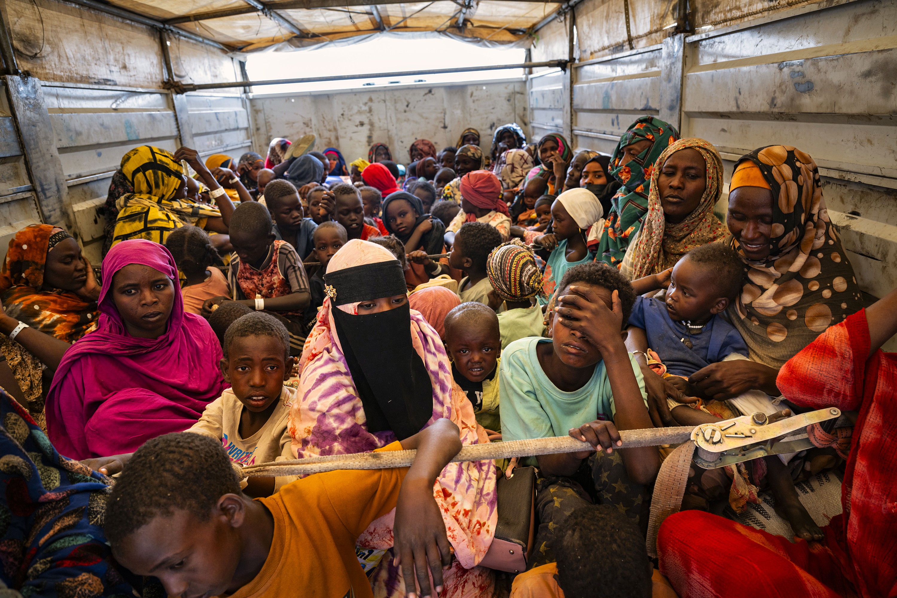 photo of interior of back of transport truck filled with women wearing brightly colored and patterned headscarves and children of all ages, with child in foreground pressing hand to face covering his eyes