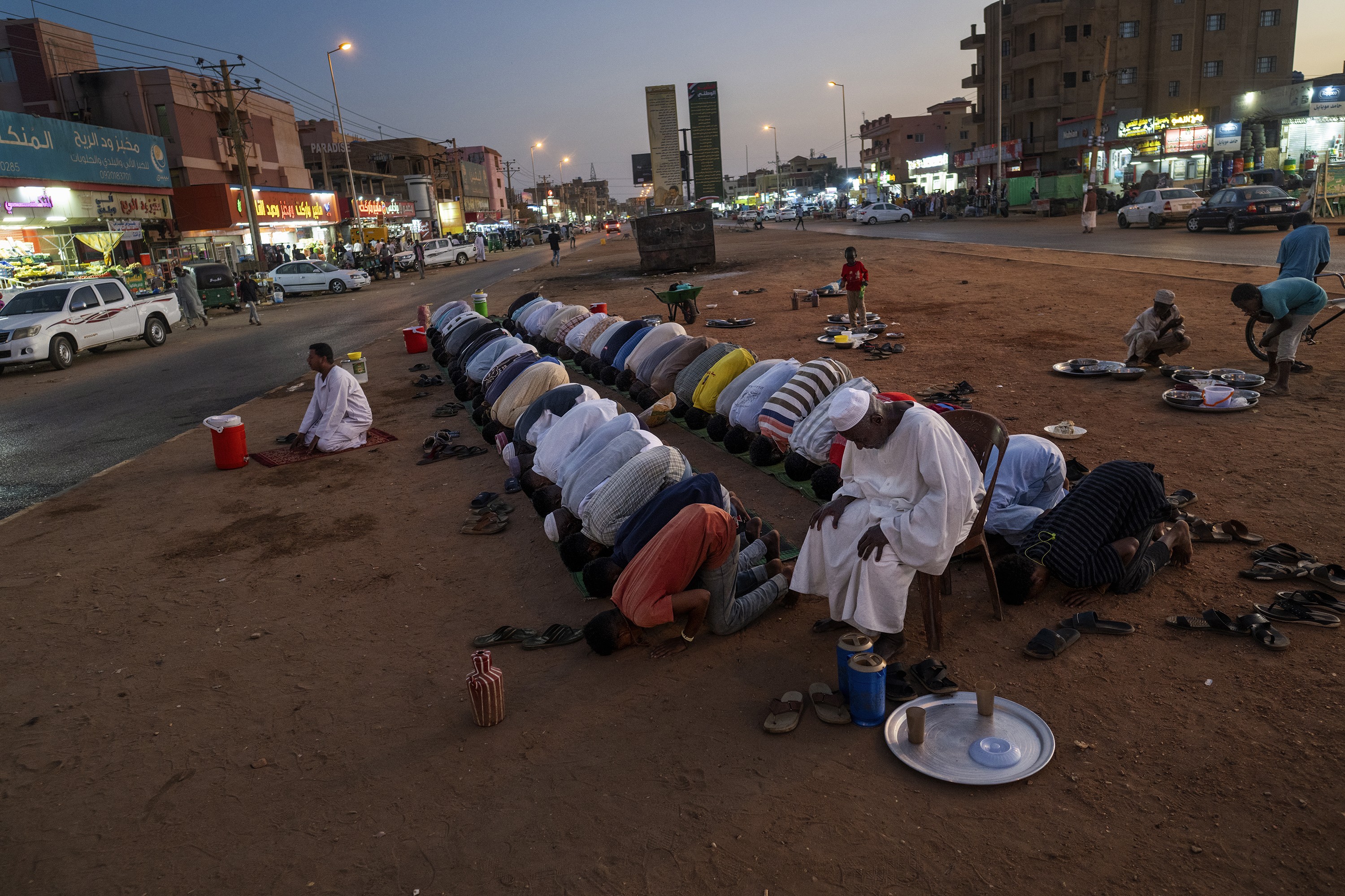 photo at dusk of two rows of men praying on wide dirt median of a street lined with shops and cars