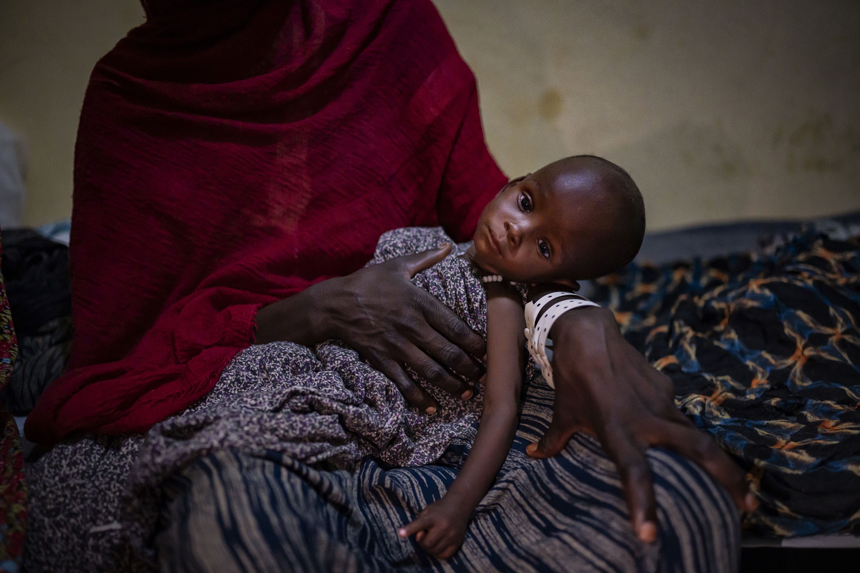 photo of woman from shoulders down wrapped in red shawl and holding emaciated baby in her lap, sitting on brightly patterned blanket