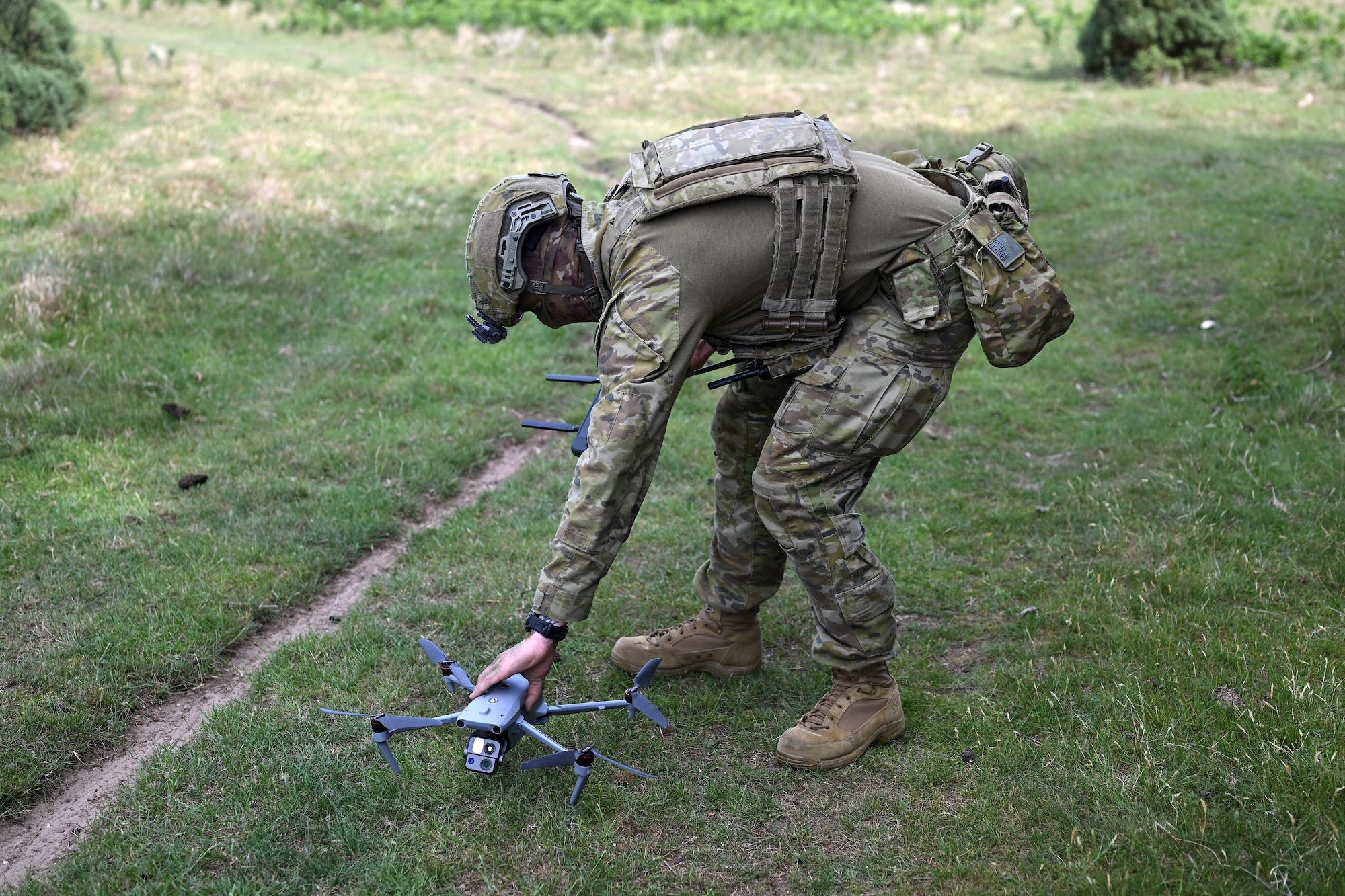 A man in camouflage gear and a helmet bends over while standing on grass to hold a small grey drone near the ground