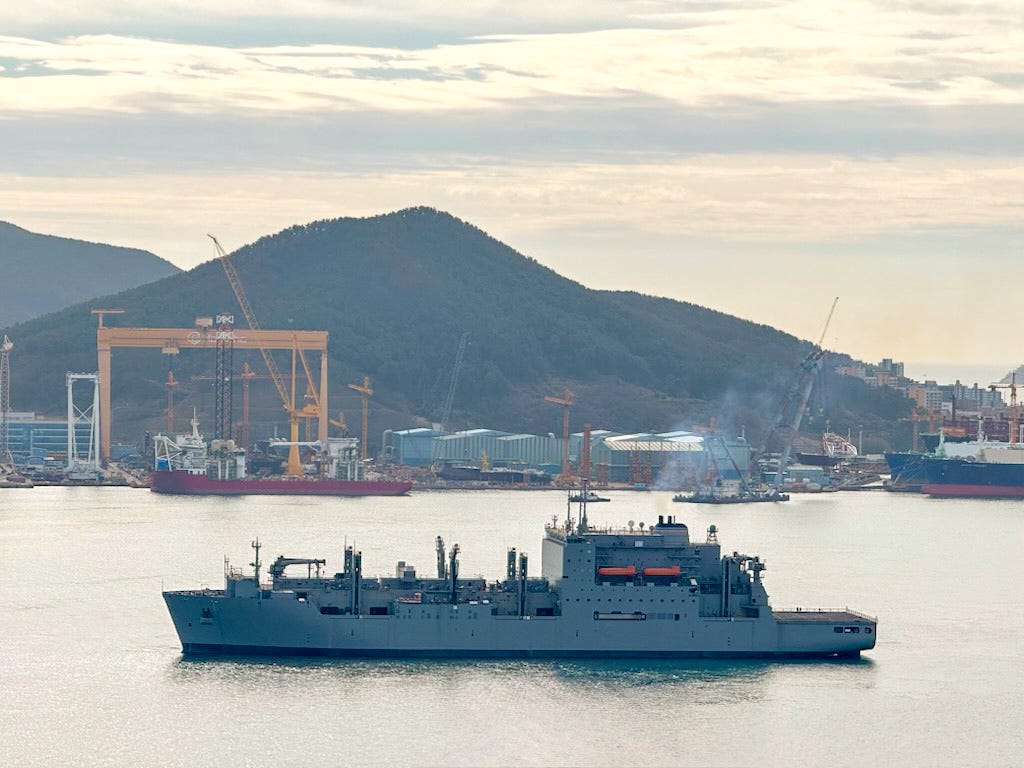 The Military Sealift Command's Lewis and Clark-class dry cargo ship USNS Wally Schirra sits in water near a South Korean shipyard. There are mountains and a cloudy sky in the background.