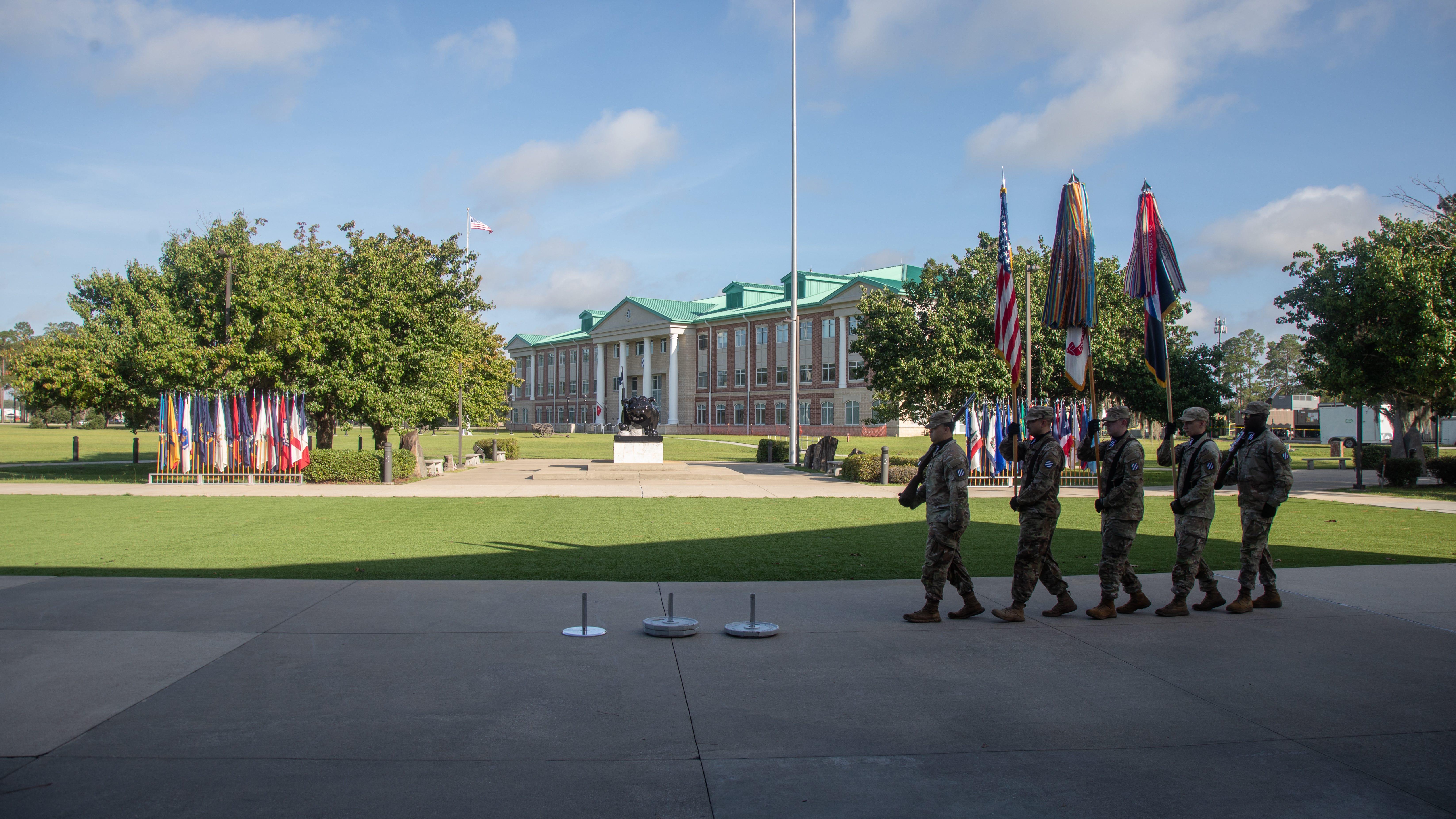 Five soldiers walk in a line on pavement with grass, trees, and a building in the background. The sky is blue with some clouds.