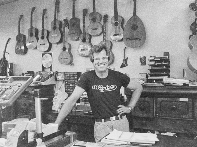 black-and-white photo of smiling man in glasses wearing black t-shirt and jeans behind the cluttered counter of music store, with guitars and instruments hung on wall in background