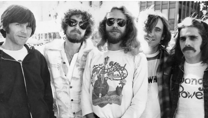 black-and-white photo of 5 men, the center one wearing sunglasses and a Westwood Music t-shirt with guitar