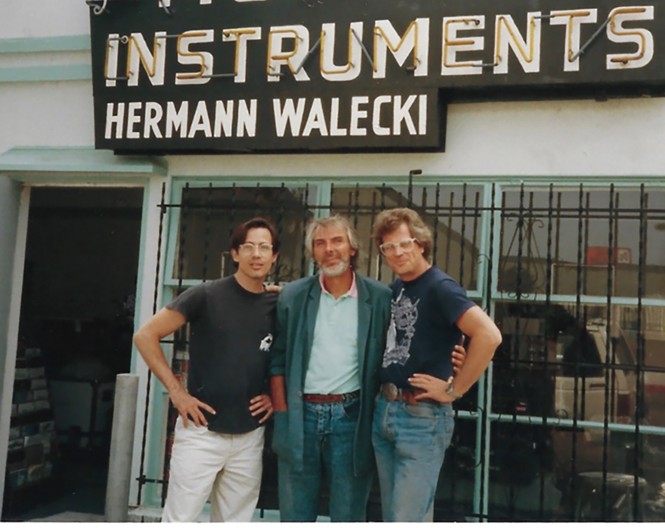 color photo of 3 men standing outside in front of store with barred display windows and large neon sign 'Westwood Musical Instruments / Hermann Walecki'