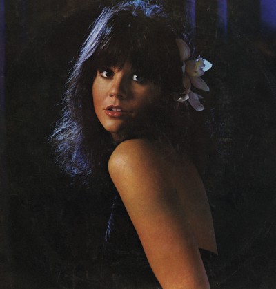studio photo of woman with long dark hair and bangs looking over her bare shoulder at camera with white flower in her hair