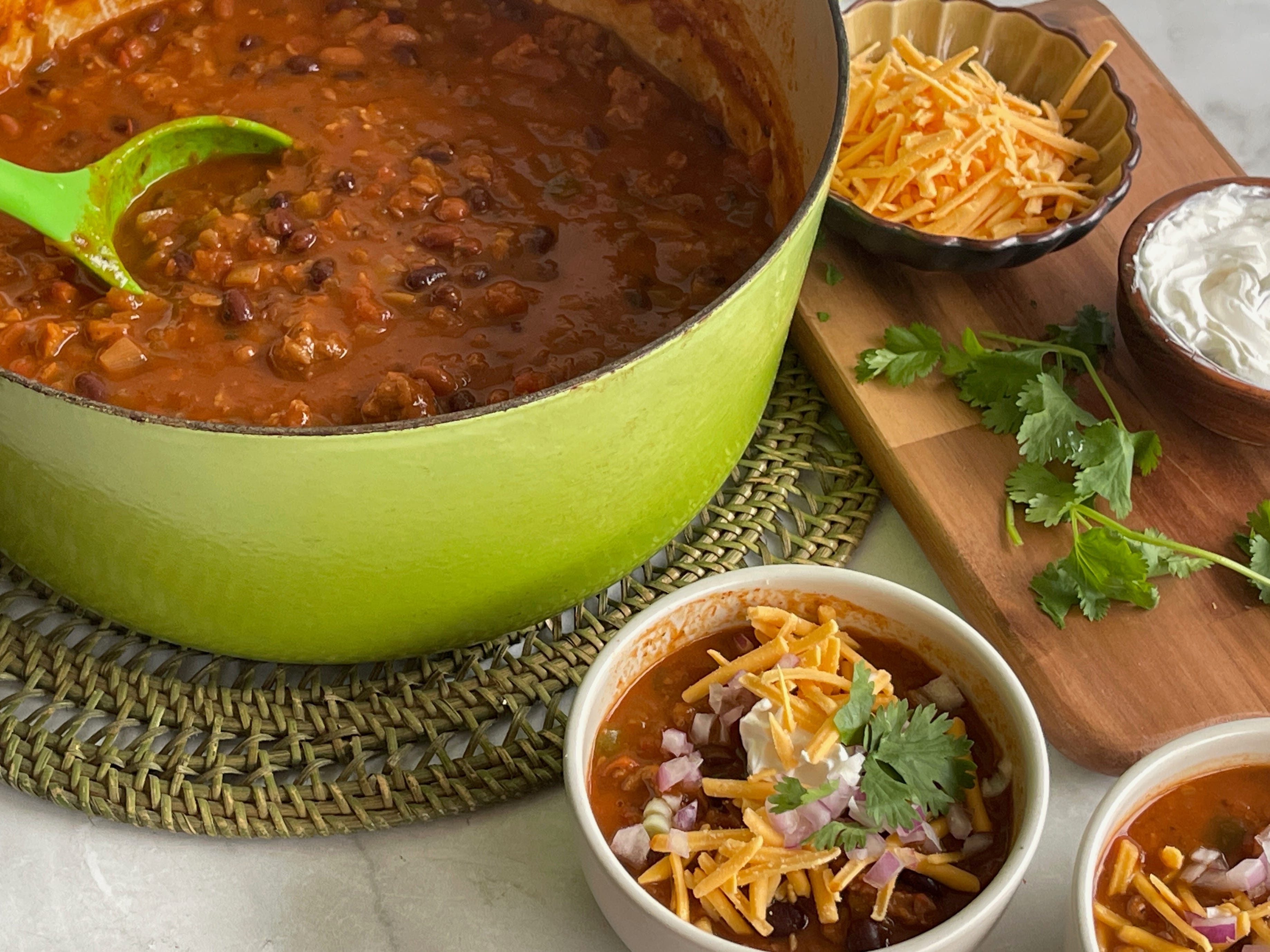 A pot of taco soup with a ladle inside, next to a bowl filled with the soup and sides of shredded cheese and sour cream.