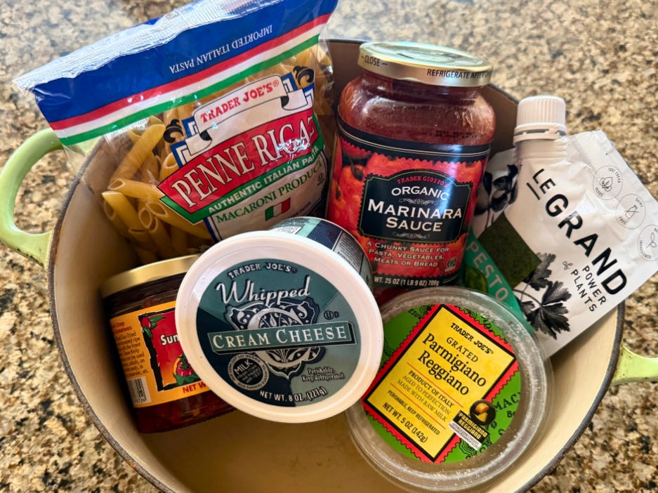 A jar of sun-dried tomatoes, a bag of penne pasta, a container of cream cheese, a jar of marinara sauce, a pouch of pesto, and a container of grated Parmigiano Reggiano in a pot on a countertop.
