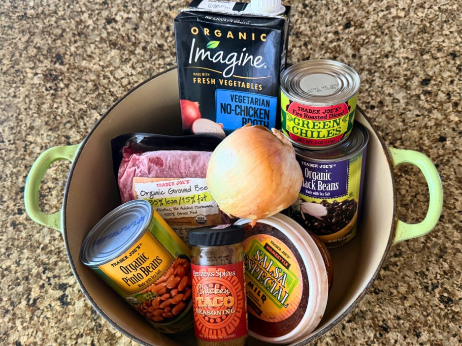 A can of pinto beans, a package of ground beef, a box of broth, a yellow onion, a jar of taco seasoning, a container of salsa, a can of green chiles, and a can of black beans in a pot on a countertop.