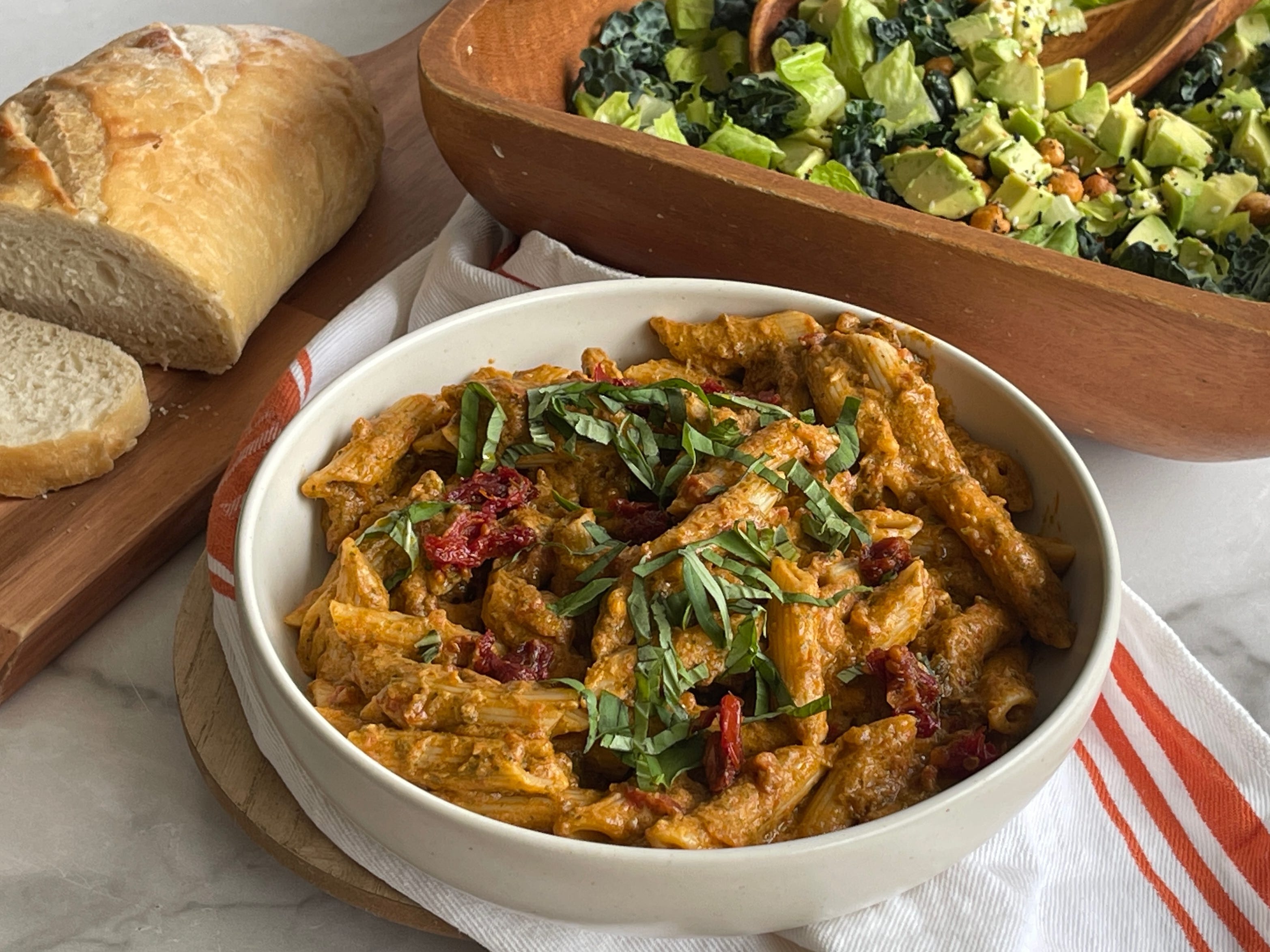 A bowl of creamy pesto pasta garnished with basil, with a loaf of bread and a salad served behind it.