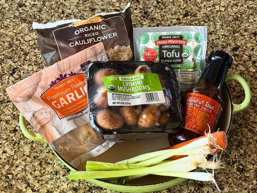 A bag of garlic, a bag of cauliflower rice, a package of mushrooms, green onions, a carrot, a bottle of stir-fry sauce, and a package of tofu in a pot on a countertop.