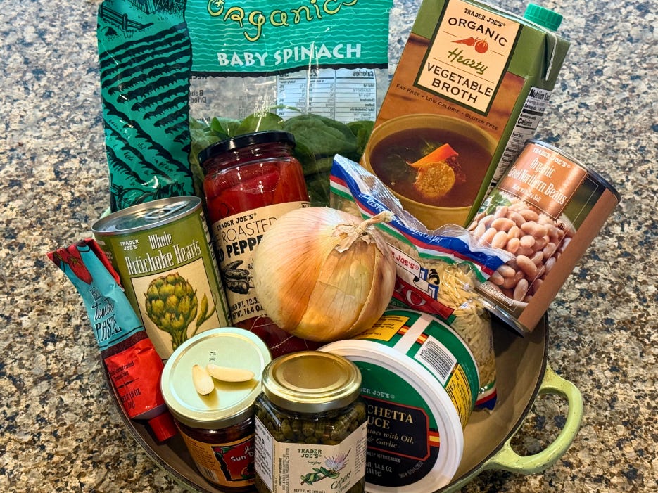 A tube of tomato pasta, a jar of artichoke hearts, a jar of sun-dried tomatoes, a bag of baby spinach, a jar of roasted red peppers, a yellow onion, a jar of capers, a container of bruschetta, a bag of orzo, a box of broth, and a can of canellini beans in a pot on a countertop.