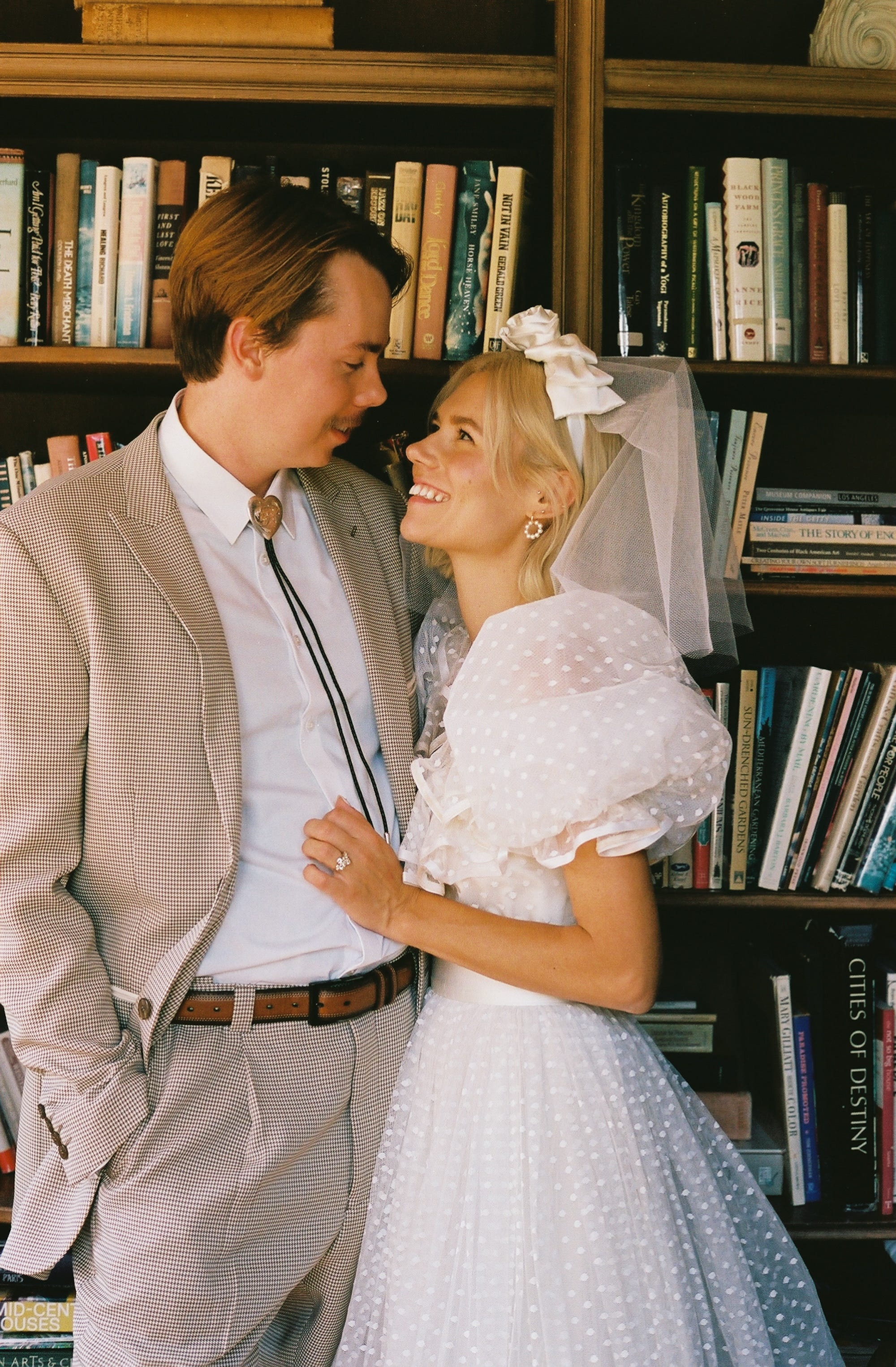 A bride and groom look at each other in front of a bookshelf covered in books.