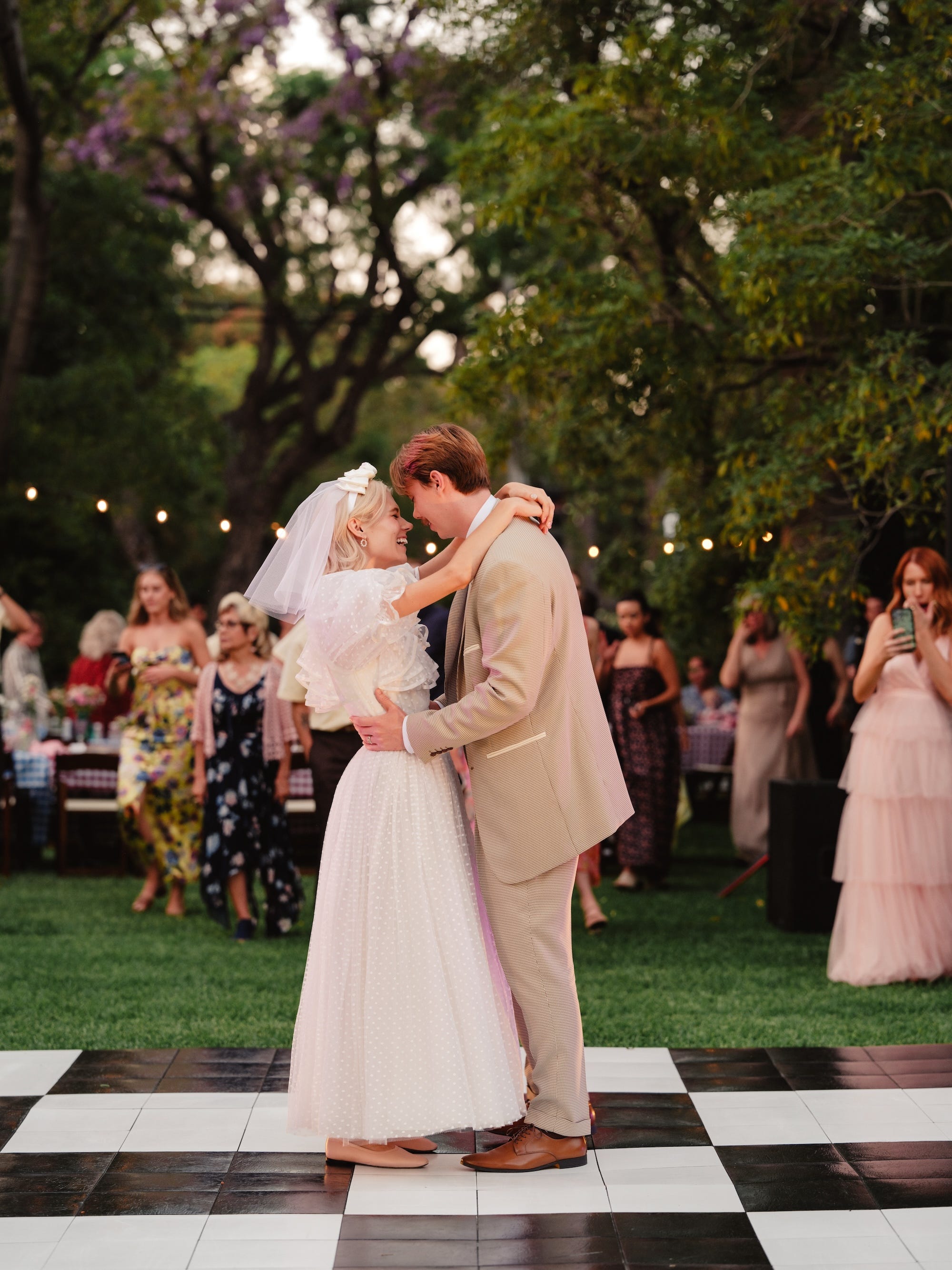 A bride and groom embrace on a checkered dancefloor at their outdoor wedding.
