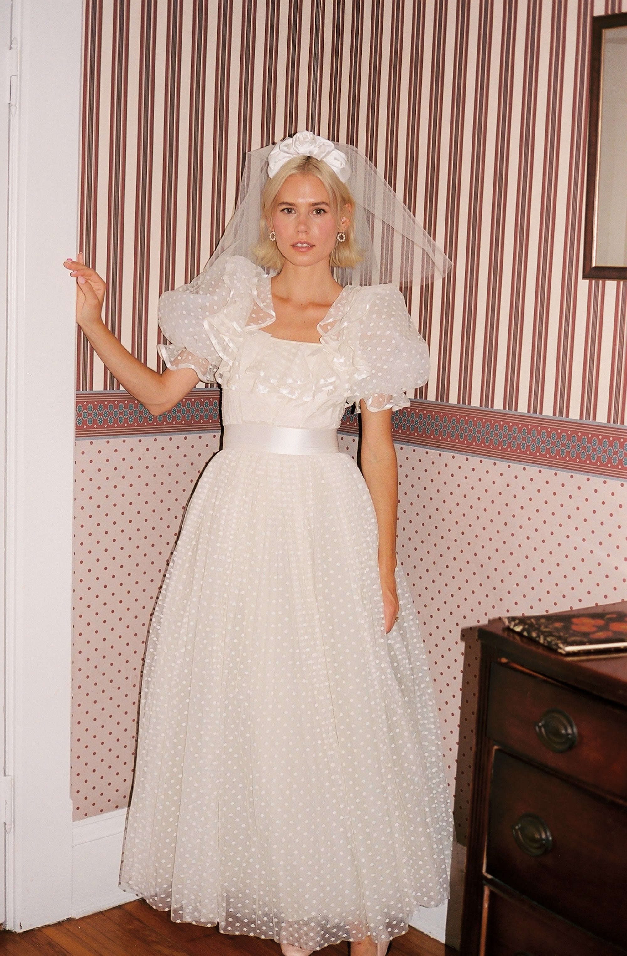 A bride stands in her wedding dress in front of a wall with patterned wallpaper.