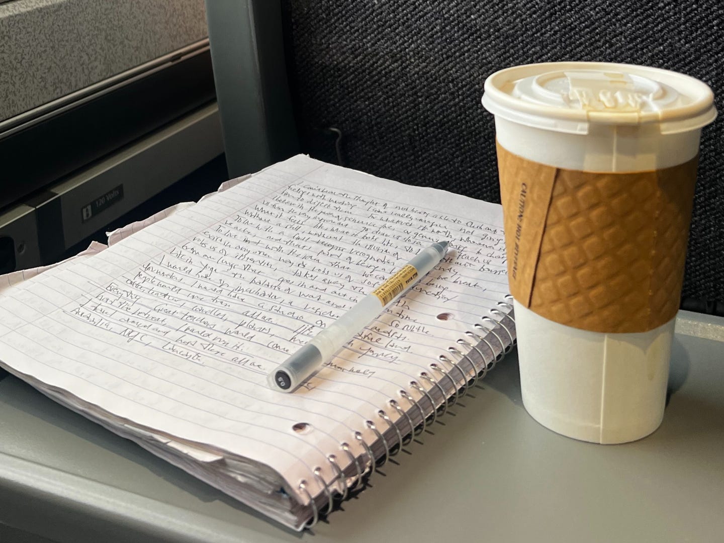 A notebook that's been written in, a pen, and a cup of coffee on a train tray table.