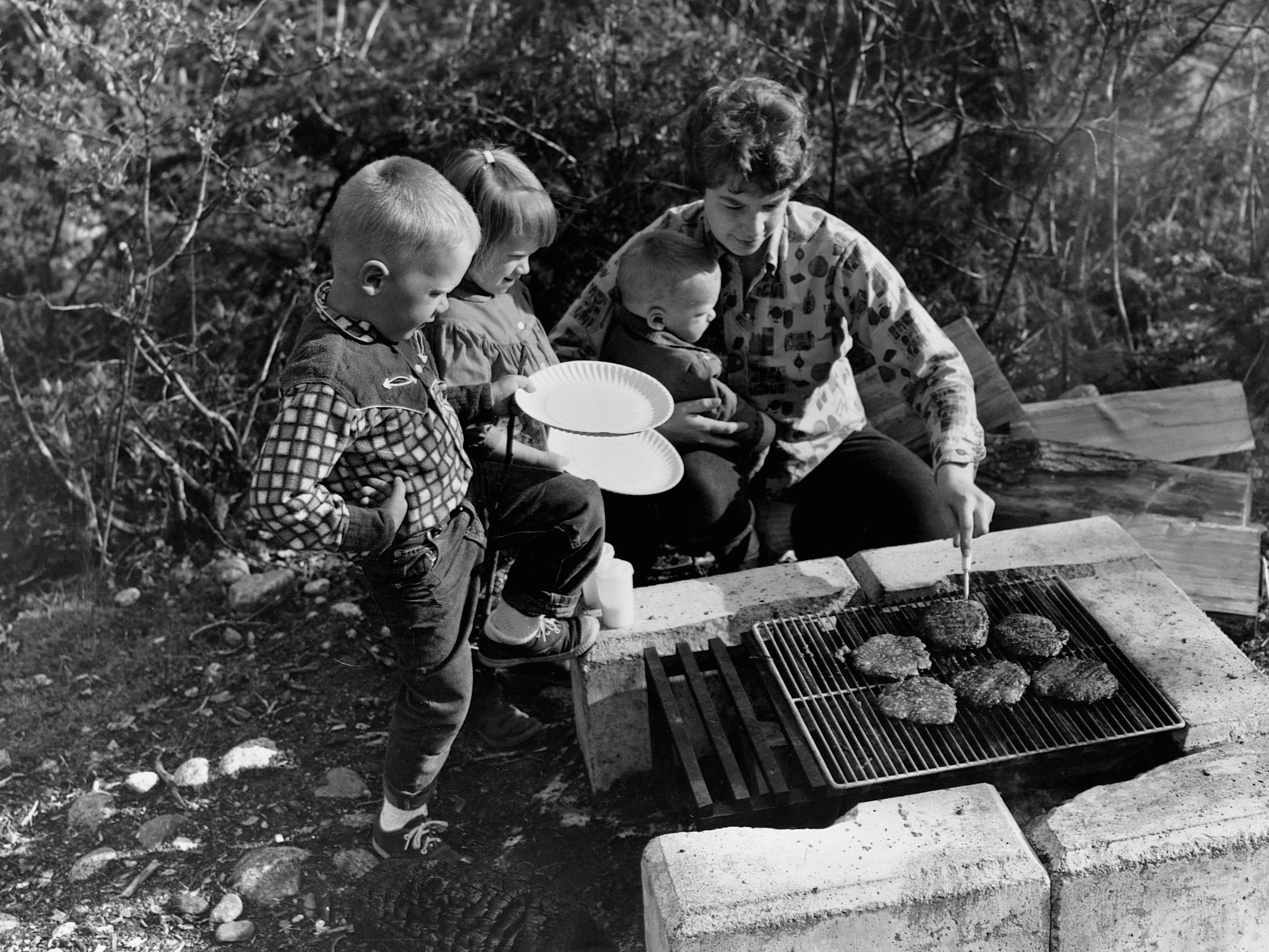 A mother grills hamburgers over a campsite grill in 1967