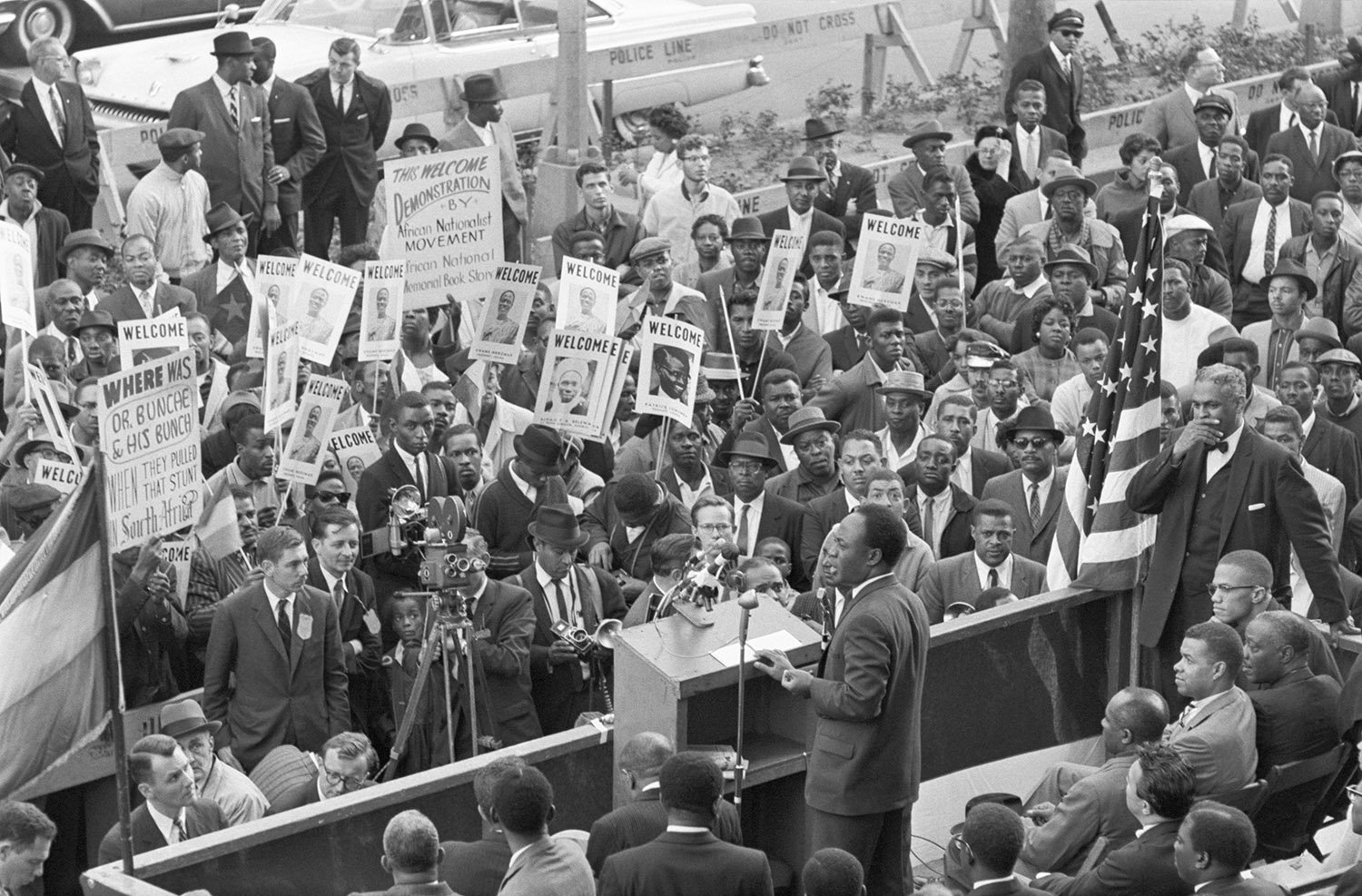 A man stands at a lectern facing a large crowed holding signs.
