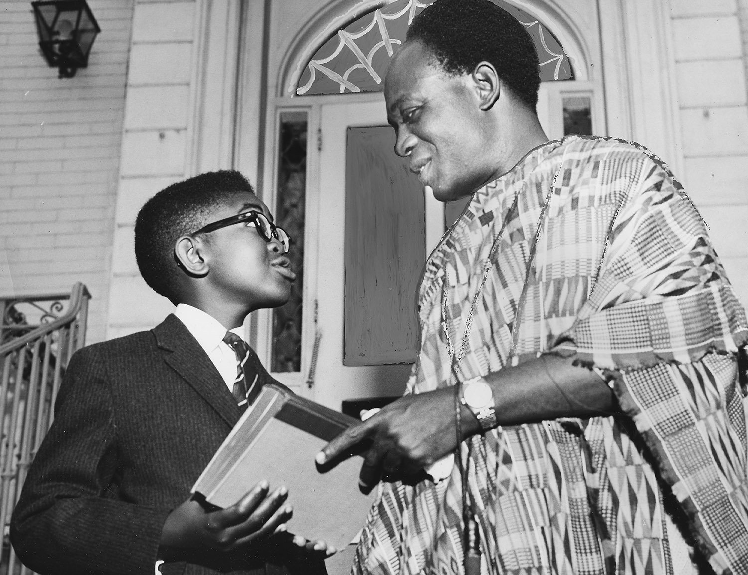 A man in a robe talks with a child in a suit and glasses who looks up at him.