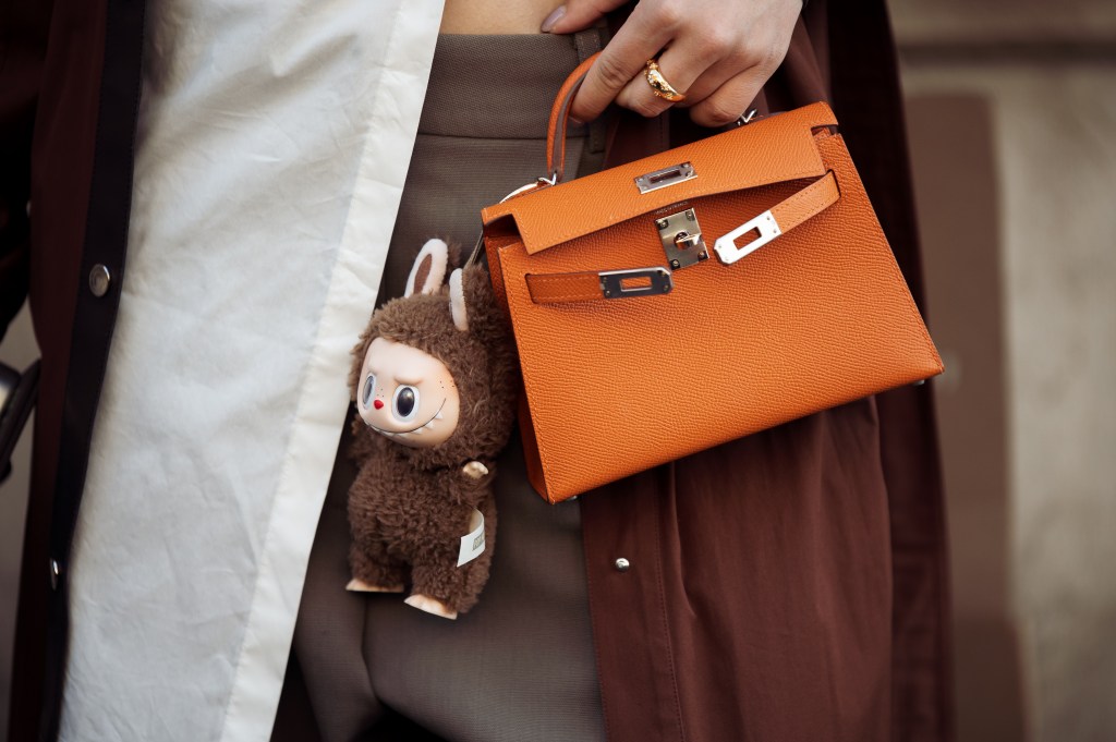 Person holding an orange Hermès bag with a brown Labubu monster charm.
