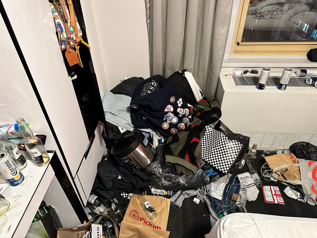 Messy hotel room with cooking equipment, clothing, and alcohol.