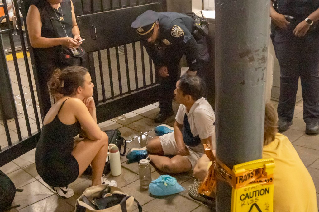 A man sits on the ground of a subway station as a police officer and bystanders look on.