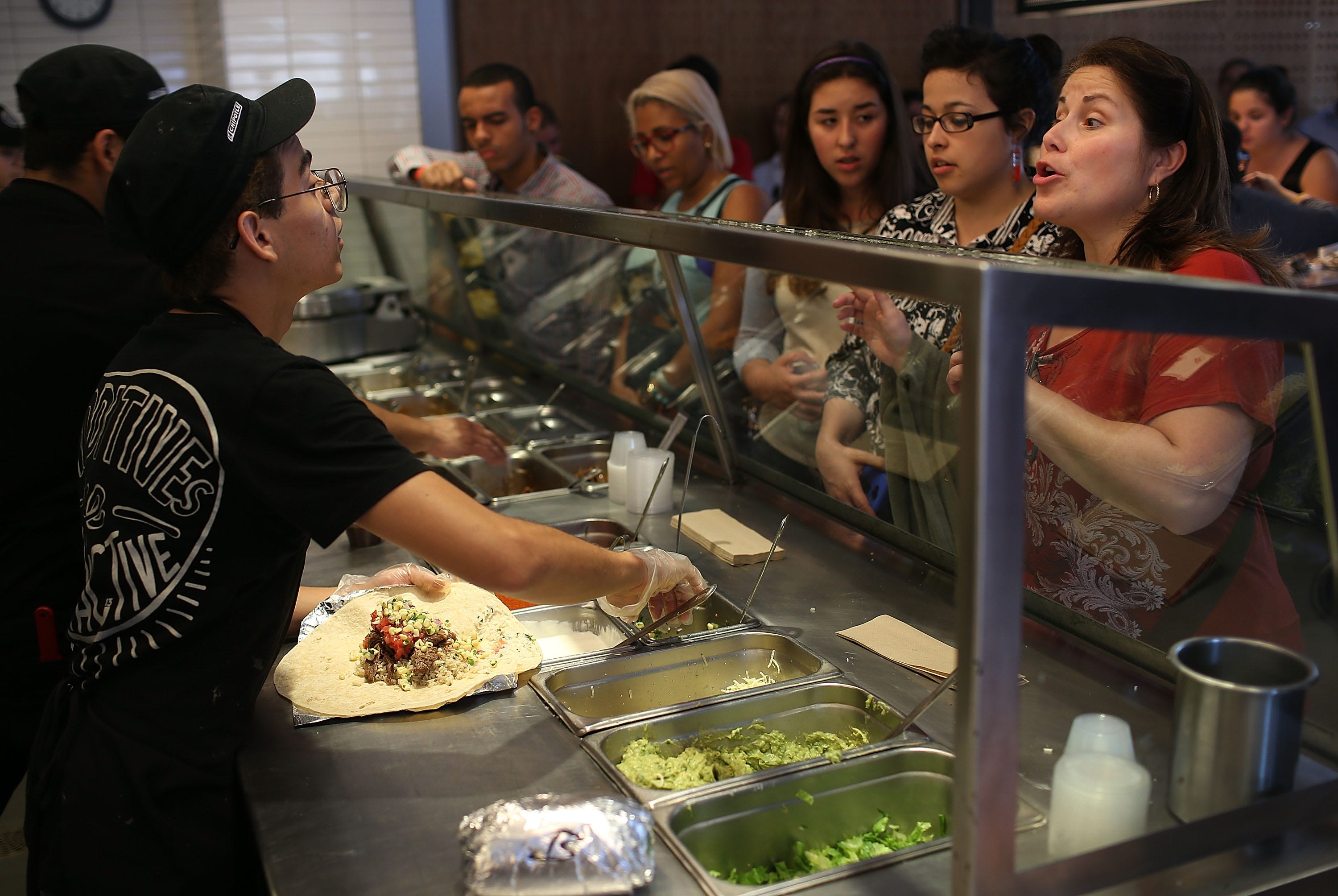 A line of customers stand behind the counter of a makeline for Chipotle.