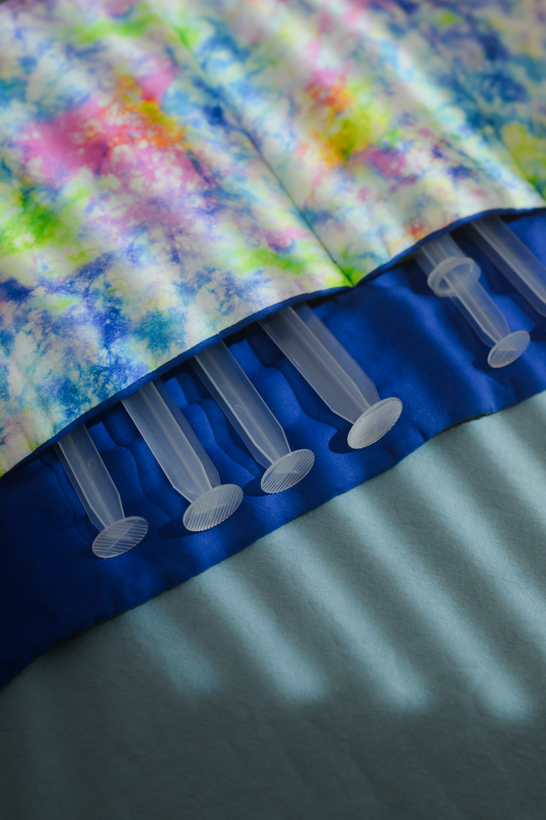 photo of brightly colored sewn cloth pouch with individual slots for syringes, with 6 syringes tucked in, the top tie-dye pastels and the bottom bright blue