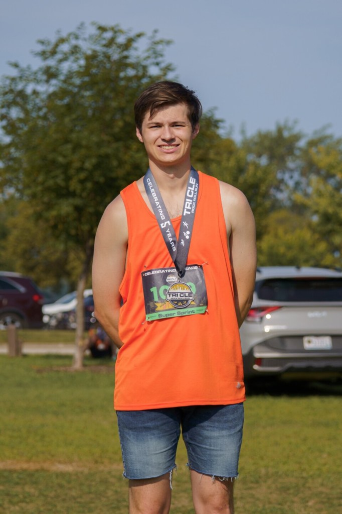 Man in orange shirt and jean shorts, wearing a medal and bib from the Tri-Cycle Rock Roll Run Super Sprint.