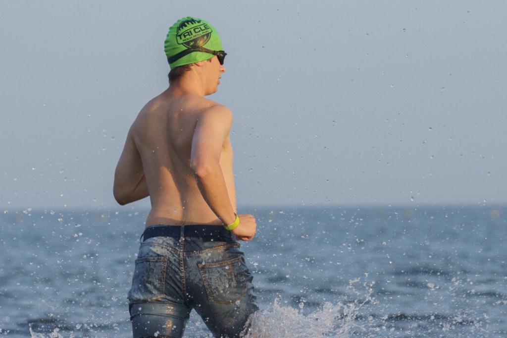Man running in the water wearing jeans and a swim cap.