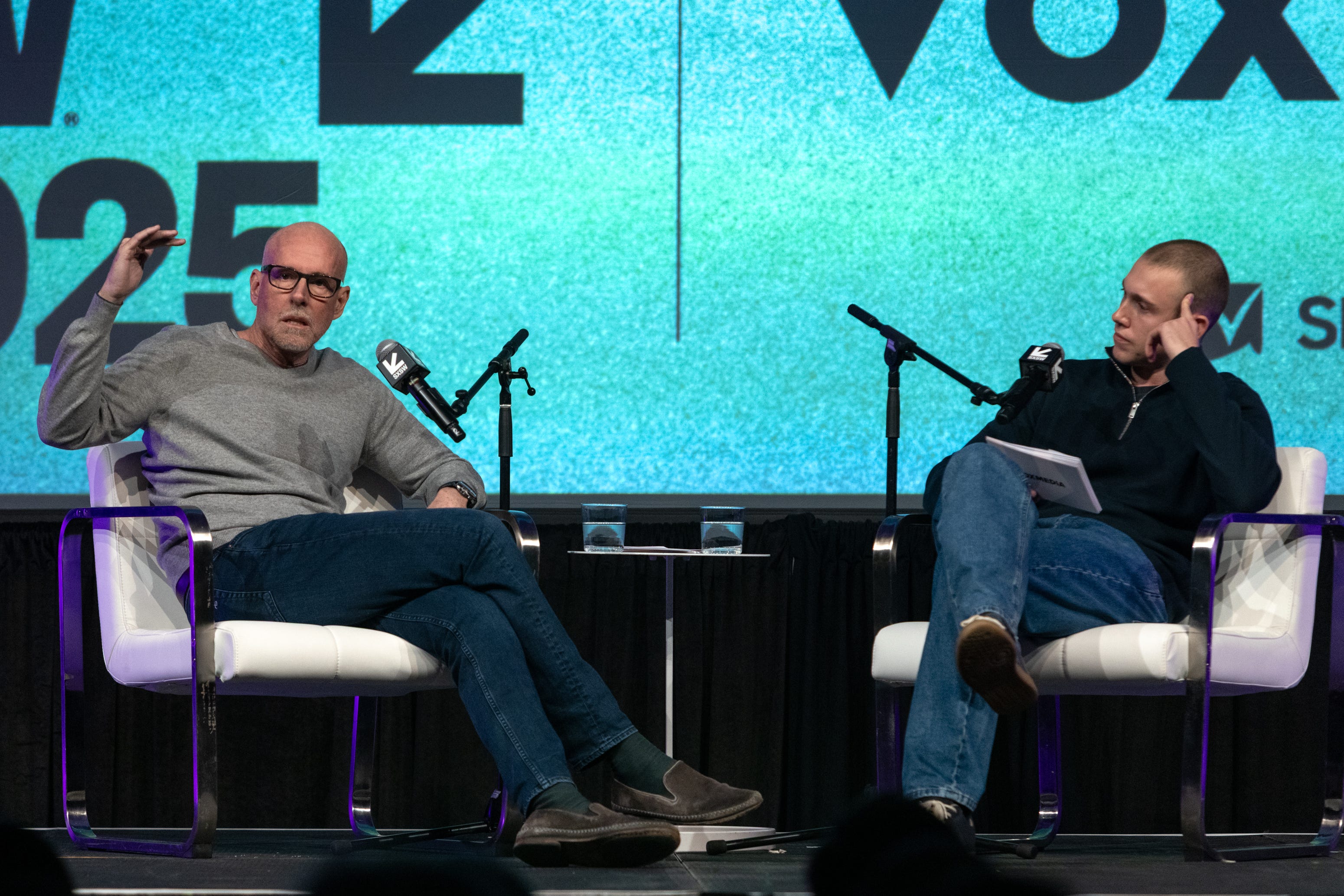 Two men sit in chairs on a stage