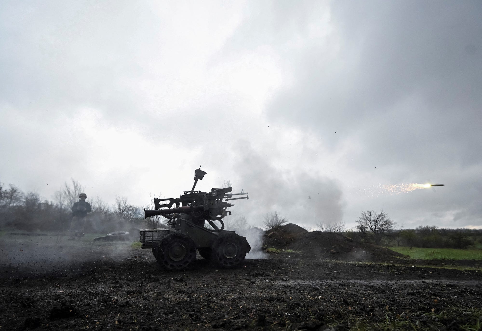 A small wheeled robot fitted with a grenade launcher rolls on a dirt ground with a grey sky and a man behind it
