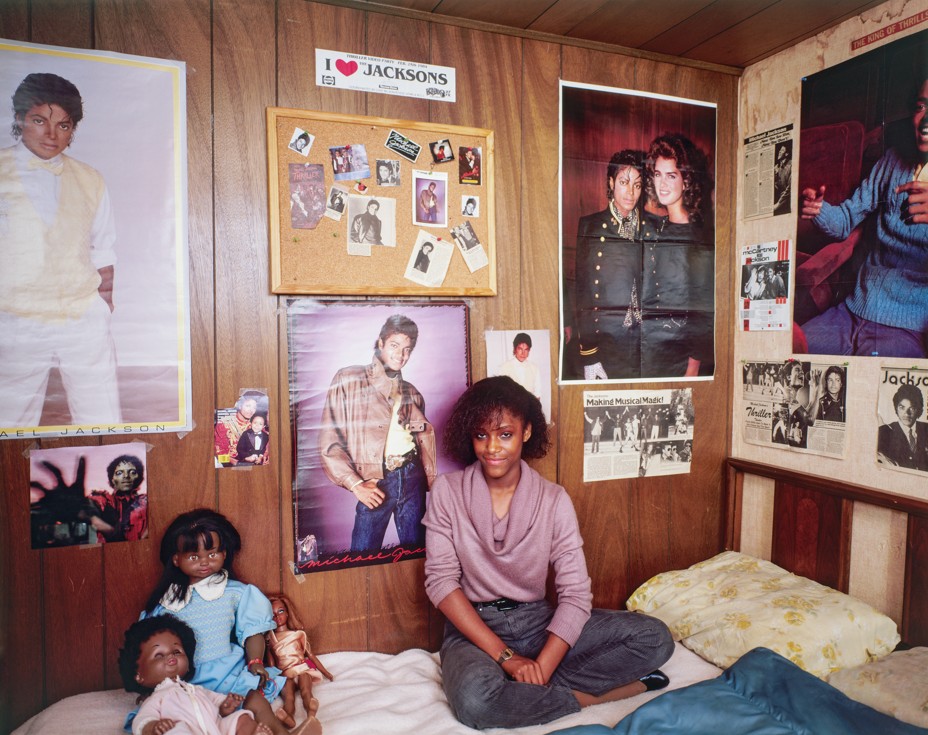 photo of girl in pink cowl-neck sweater and jeans sitting cross-legged on bed leaning against wood-paneled wall with many posters of Michael Jackson, a bulletin board, newspaper clippings, and a bumper sticker with 
