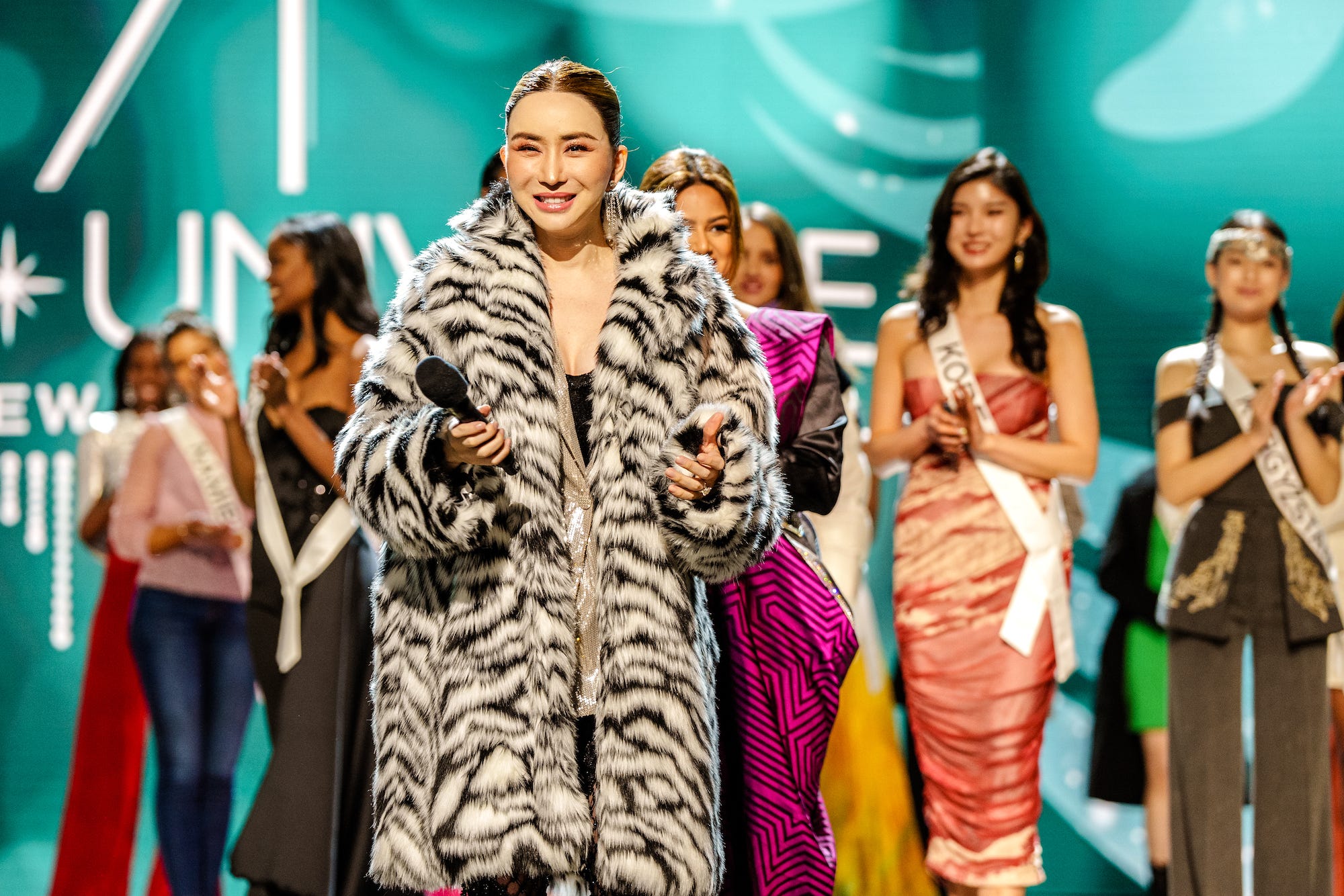 Anne Jakkaphong Jakrajutatip on the Miss Universe stage wearing a fur coat and holding a microphone.
