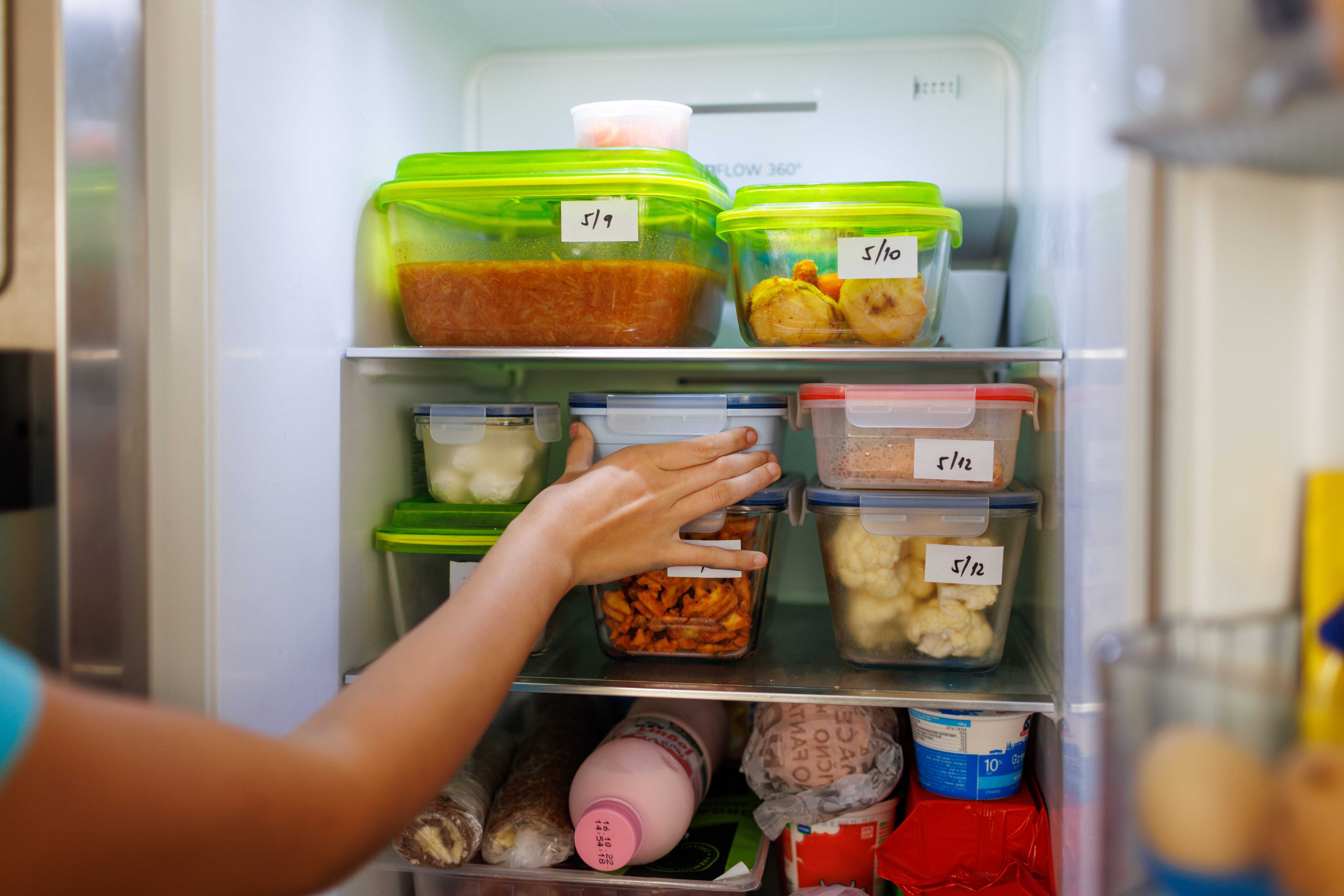 An arm reaches into a fridge with labeled containers of food.