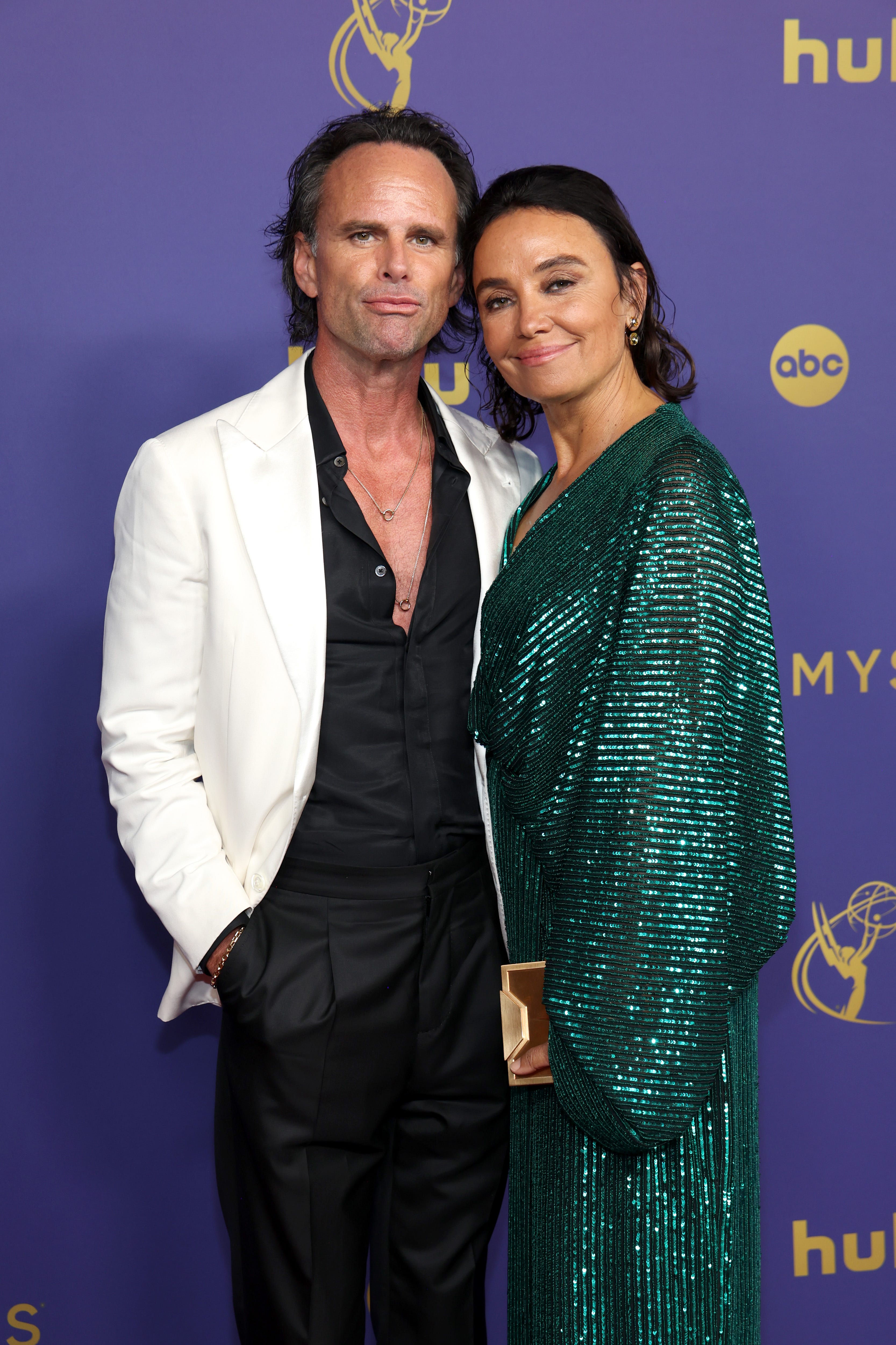 A man and a woman at an event. On the left, the man has long swept-back black hair. He's wearing a white blazer over an open-collared black shirt and black trousers. On the right, the woman also has her black hair swept back, and is wearing a glittery green dress. They're standing against a purple backdrop with gold logos for Hulu, ABC, and the Emmys on it.