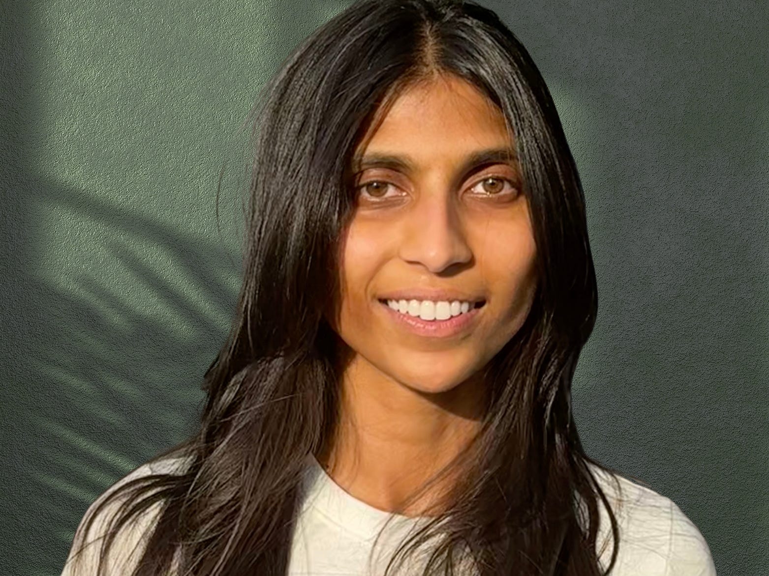 A headshot of Maya Bakhai in front of a green wall with the shadow of a tree behind her.
