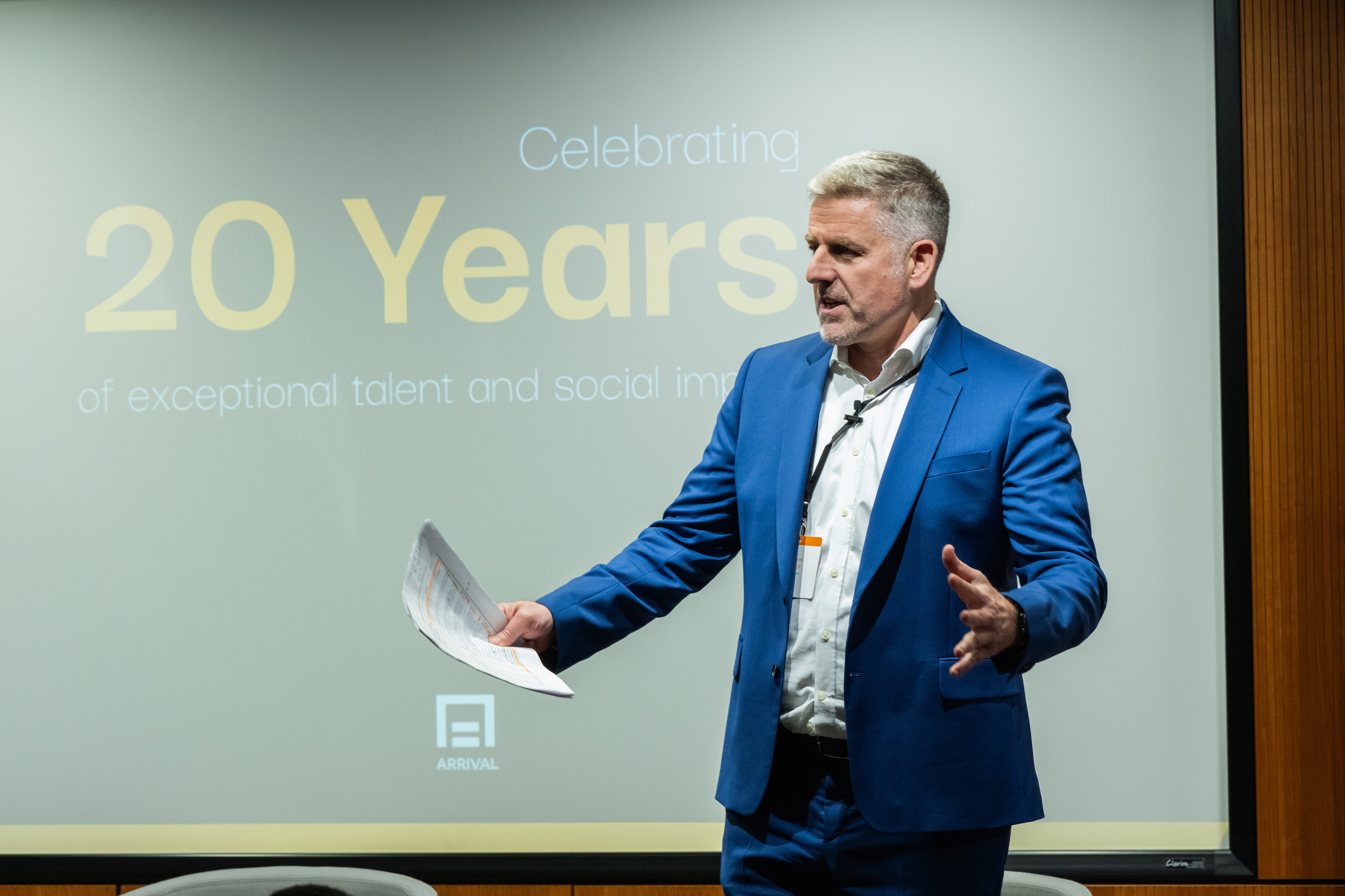 A man with grey hair, in a blue suit, holding a piece of paper and standing in front of a screen that says, "Celebrating 20 years."