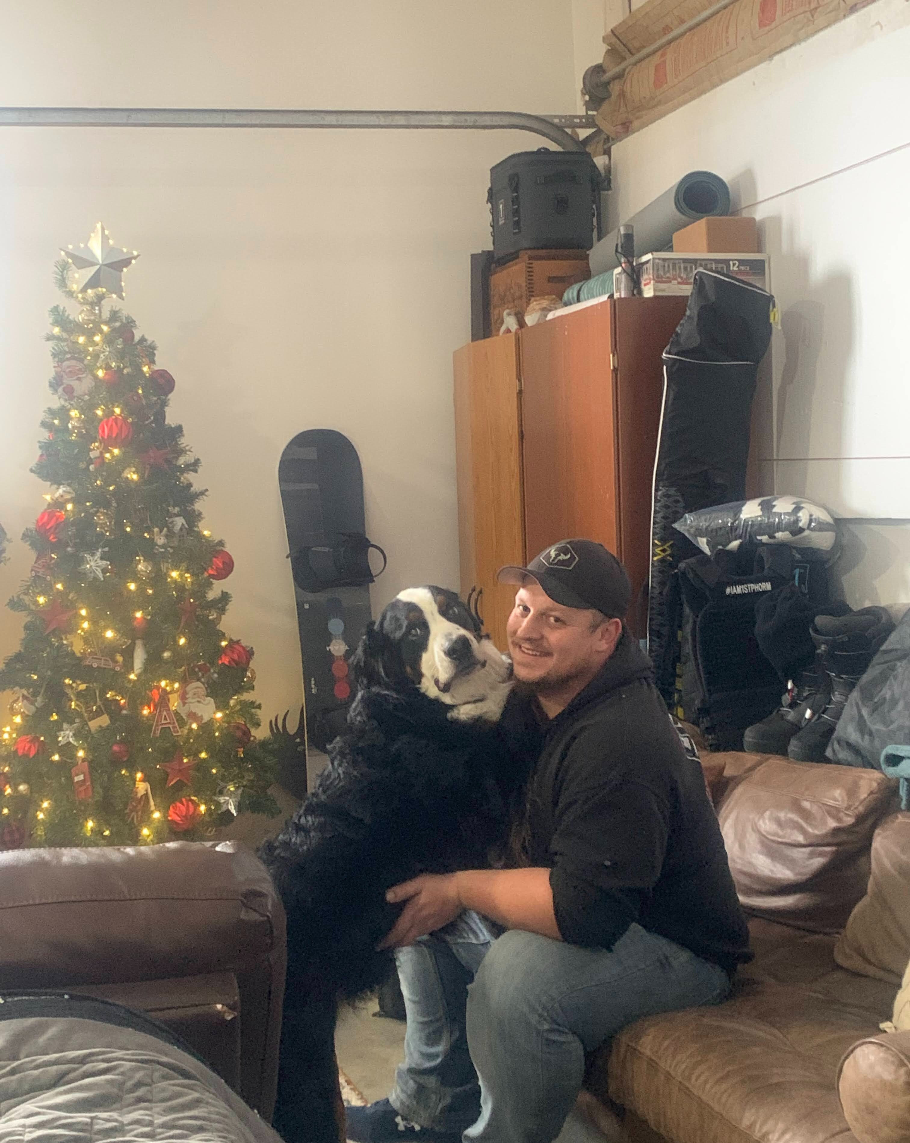 man and dog smiling for camera inside garage with fake small xmas tree in background