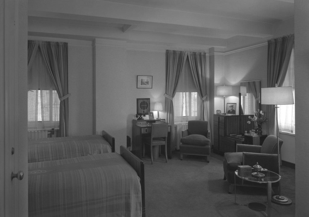 Black and white photo of a hotel room with twin beds, a desk, chairs, and a radio.