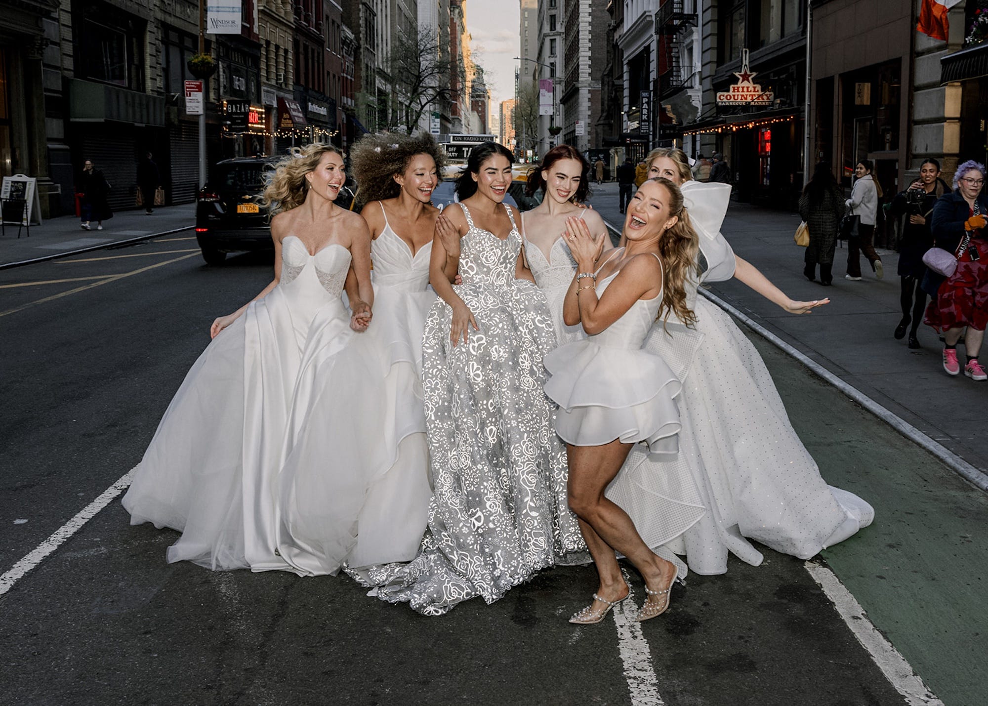 Hayley Paige claps in front of a group of women wearing wedding dresses on a street.